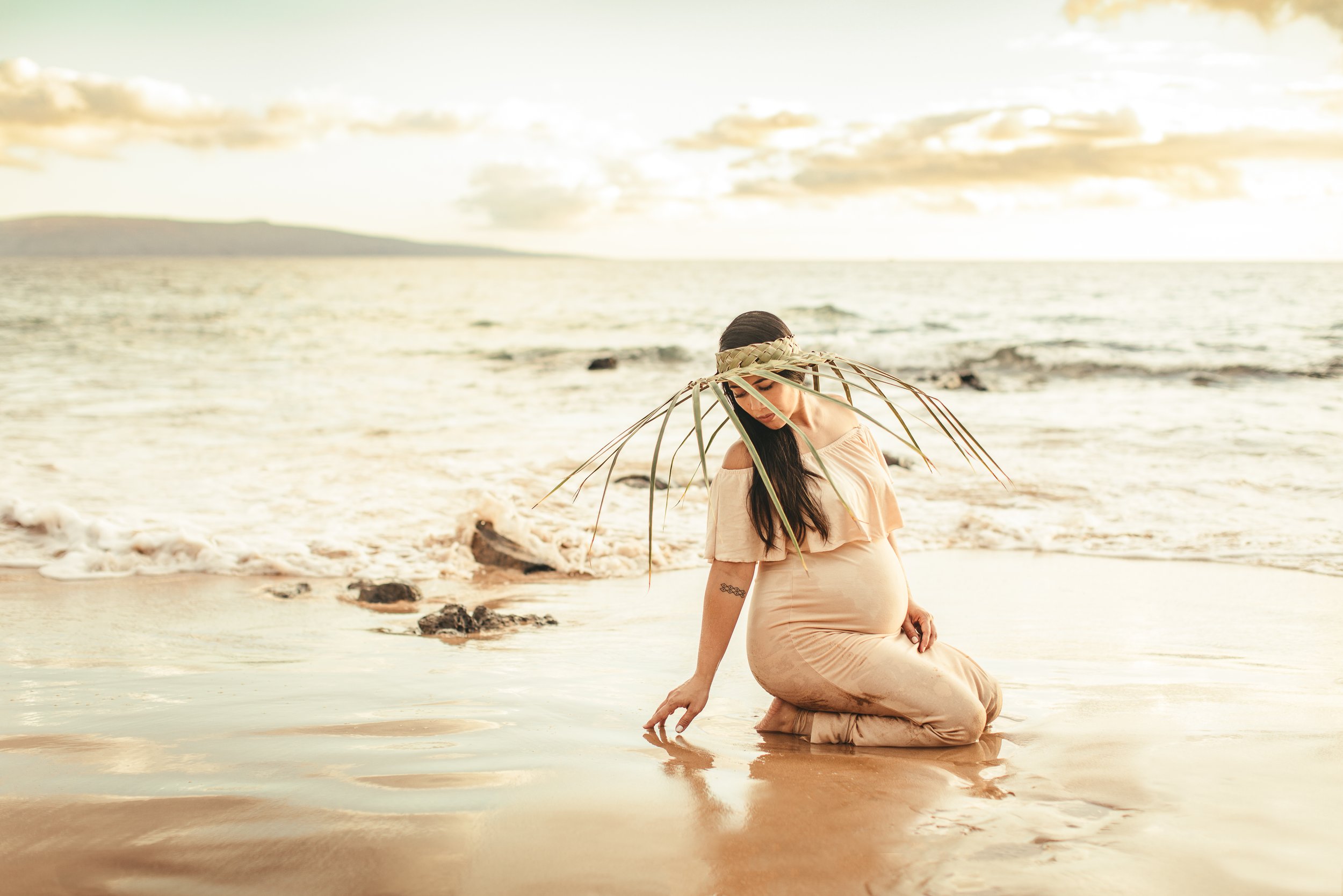 pregnant hawaiian woman kneeling on beach with palm straw hat in maui hawaii