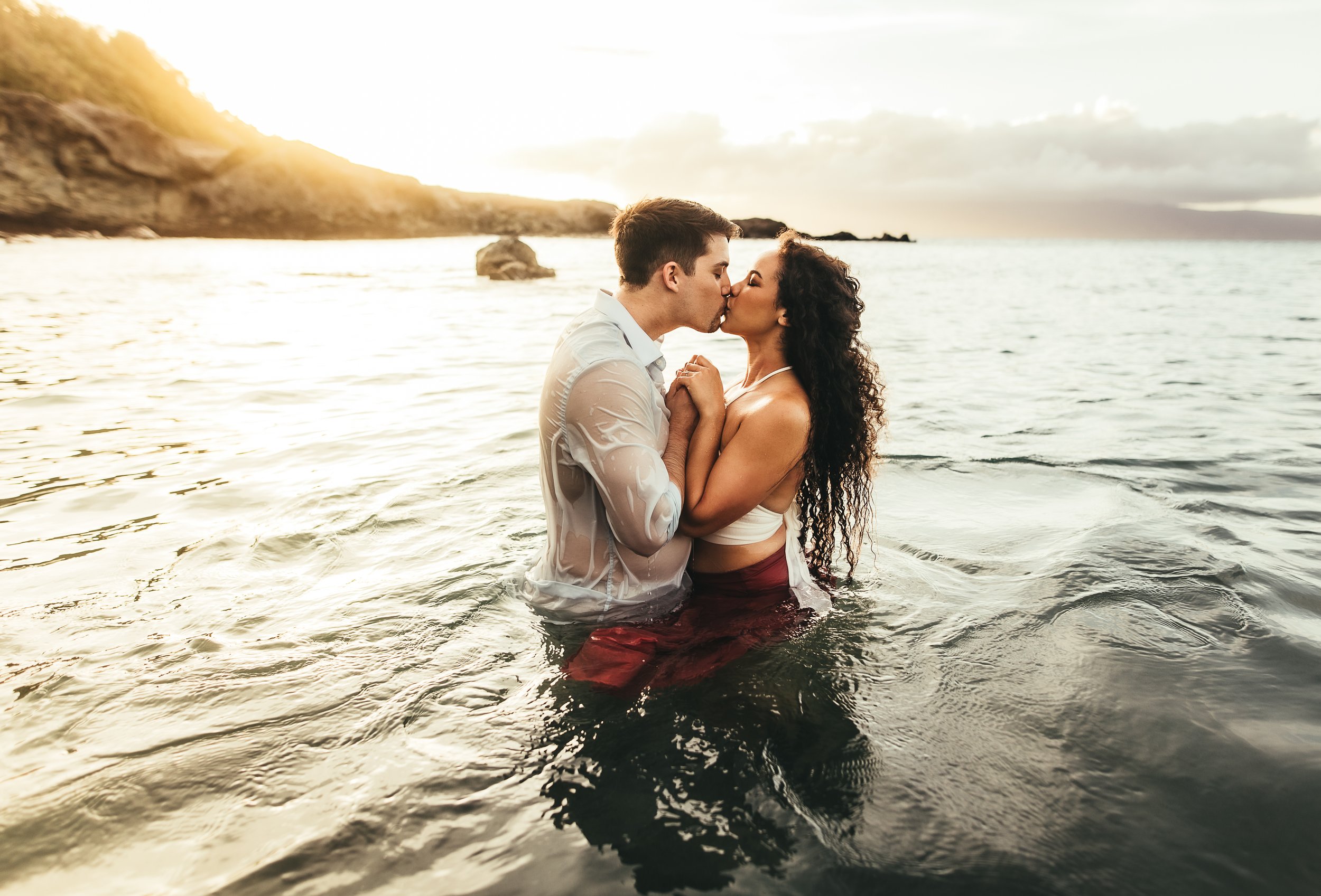 white man and african american woman standing in ocean while holding hands and kissing each other in maui hawaii
