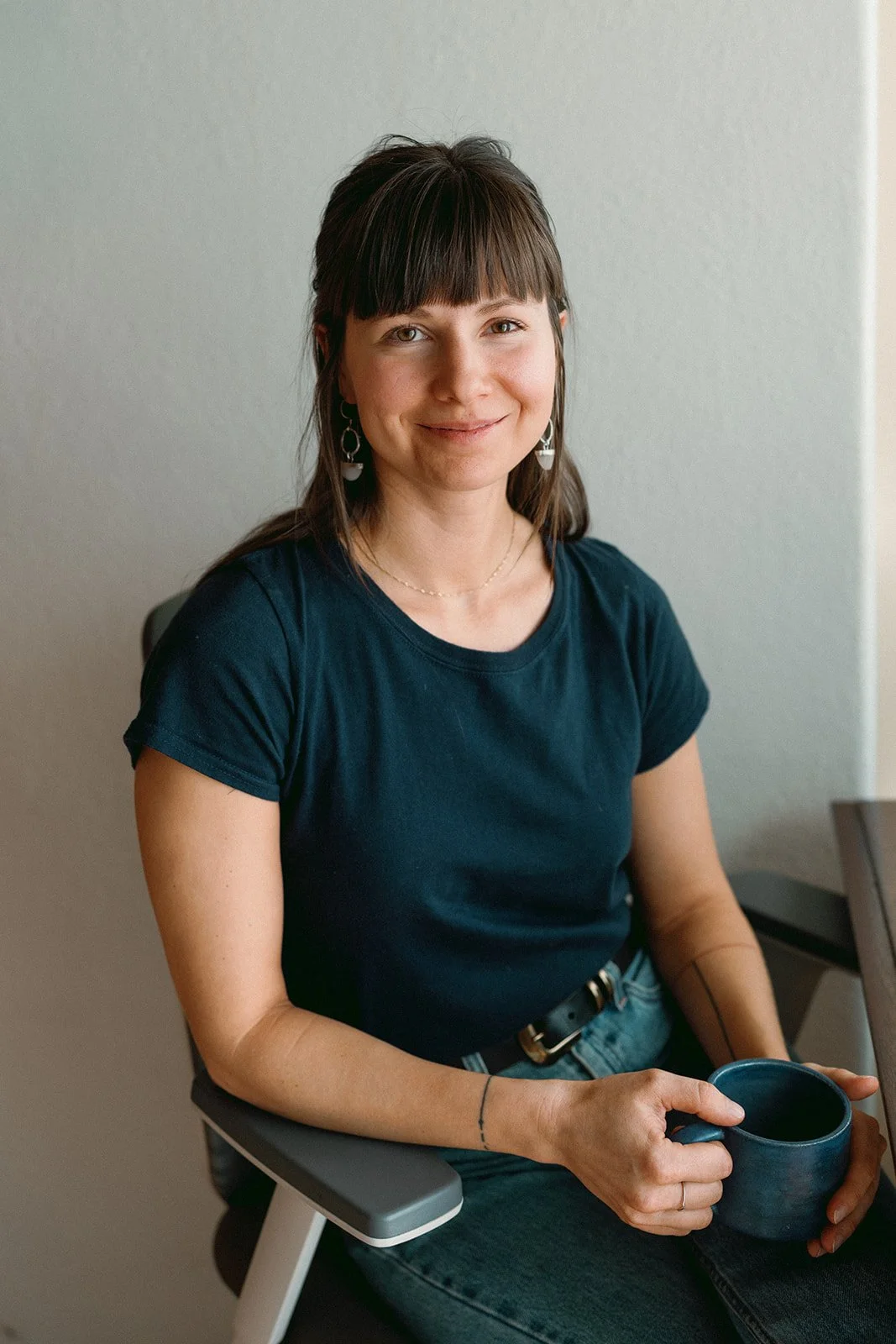 A woman with dark hair and bangs, wearing a dark teal t-shirt and earrings, sitting in a gray chair and holding a dark blue mug, smiling at the camera.