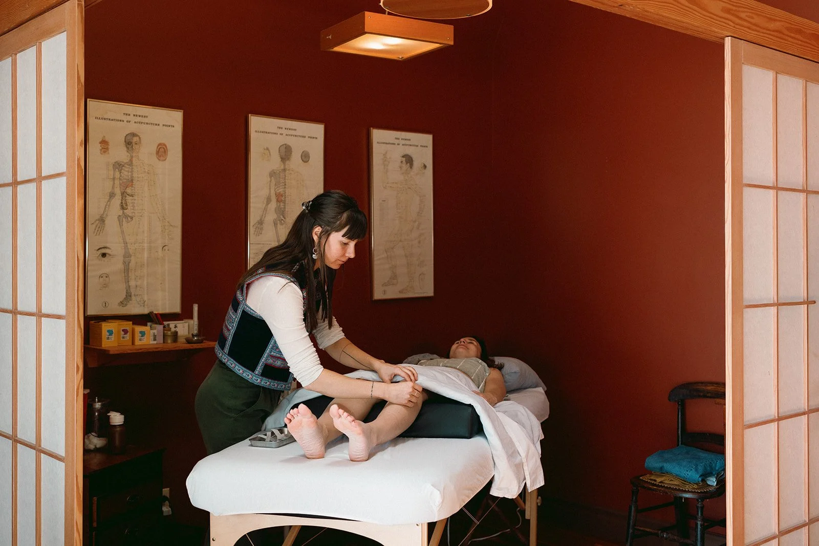 A woman lies on a massage table with her legs raised, while another woman in traditional attire performs acupuncture in a room with red walls and anatomical posters.