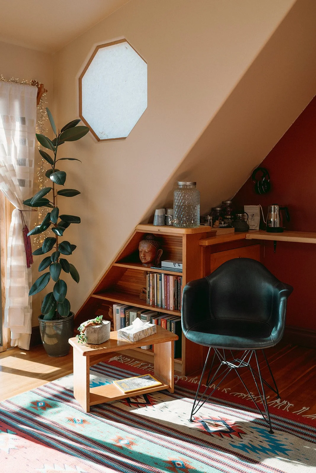 A cozy living space corner with a black chair, wooden bookshelves, a potted plant, a small coffee table, and a colorful patterned rug. Includes a skylight and window with curtains.