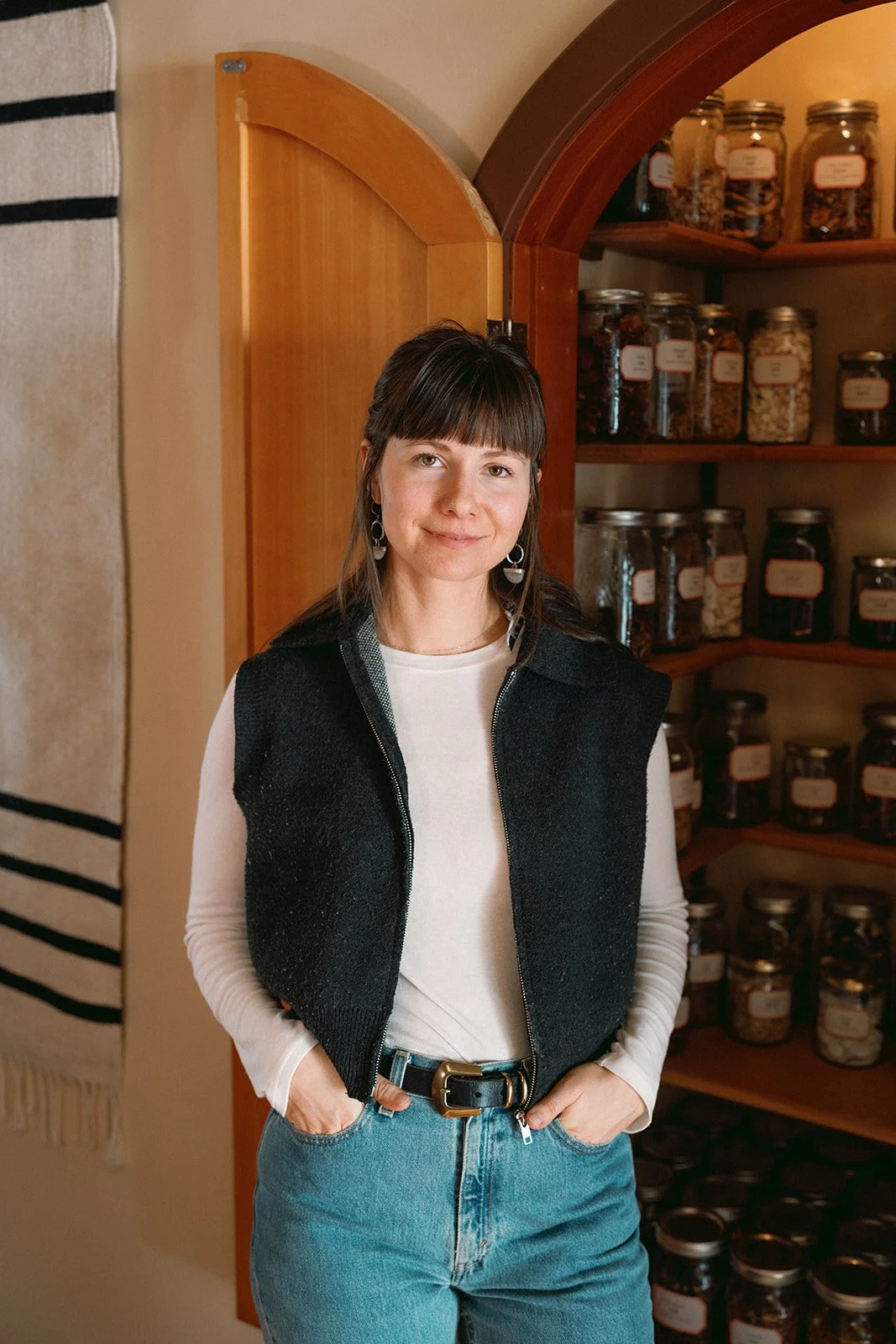 A woman standing indoors in front of an apothecary, wearing a white long-sleeve shirt, a black vest, and blue jeans with a black belt.