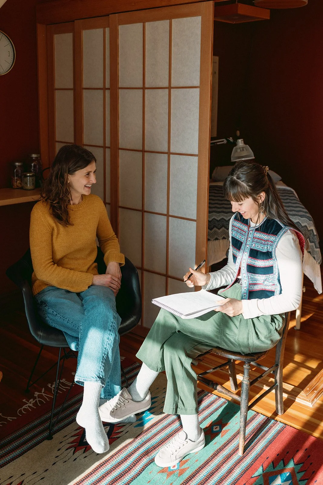 Two women sitting and smiling during a conversation in a cozy room with a wooden floor, colorful patterned rug, and a room divider.