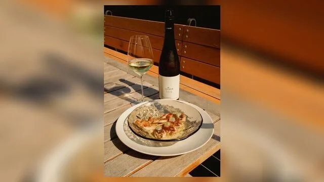 A dinner setting with a plate of pizza, a glass of white wine, and a bottle of wine on a wooden table outdoors.