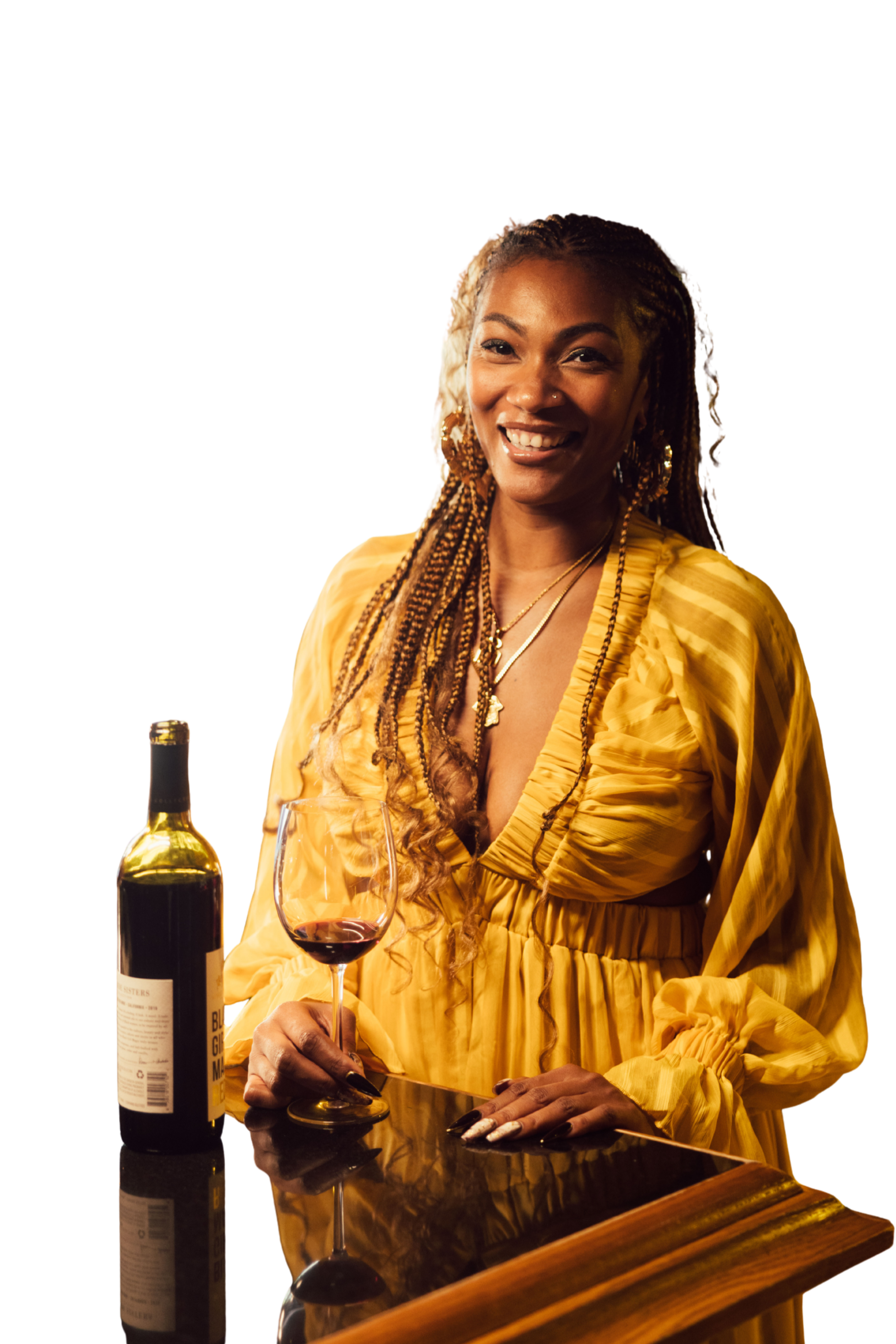 A woman with braided hair, gold earrings, and a gold necklace smiling at the camera, holding a glass of red wine in one hand, with a bottle of red wine on the bar in front of her.