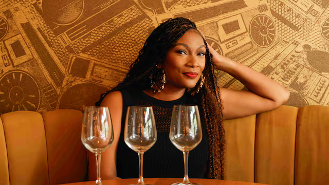 A woman with long, braided hair sits at a table in a restaurant with three empty wine glasses in front of her, wearing a black dress and earrings, with a patterned wall in the background.