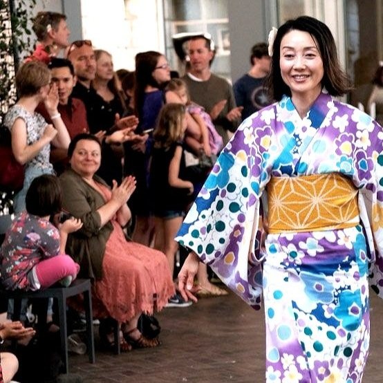 A woman wearing a purple, blue, and white floral yukata with a yellow obi walks in front of an audience at a cultural event, with people clapping and enjoying the performance.