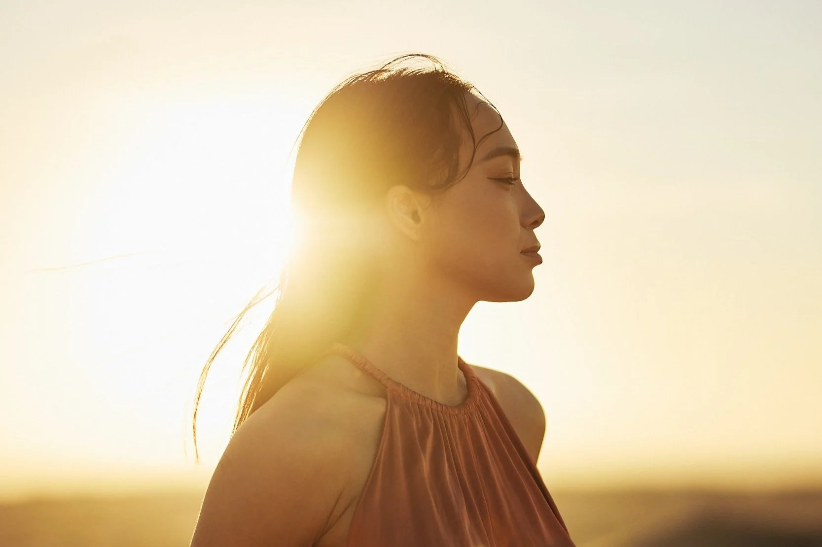 A woman in a sleeveless top stands in profile with eyes closed, in front of a bright sunlight background during sunset or sunrise.