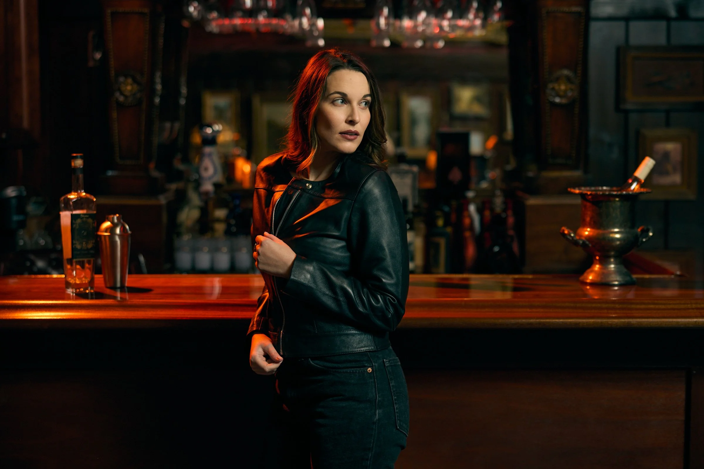 A woman with dark hair and a black leather jacket standing at a bar, looking to her left with a thoughtful expression. The background includes bottles and decorative items, creating a dimly lit, vintage atmosphere.