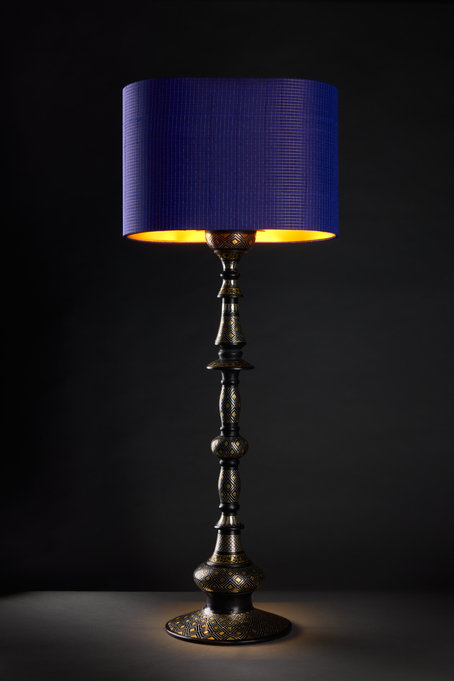 A tall decorative lamp with a black, gold, and white patterned base and a large, cylindrical dark blue lampshade emitting a warm yellow light.
