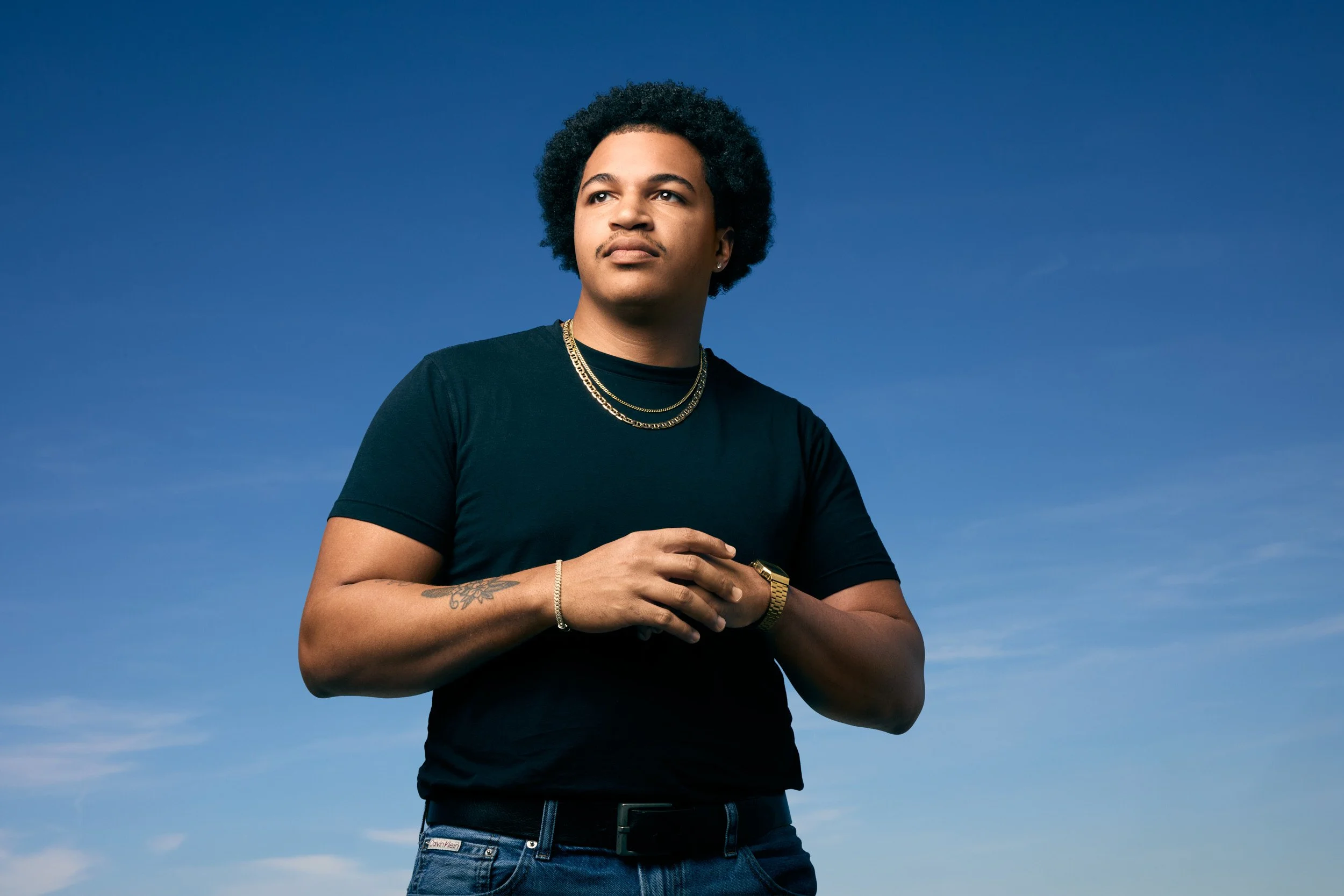 A young man with curly hair and tattoos wearing a black t-shirt and gold jewelry, standing outdoors against a clear blue sky.