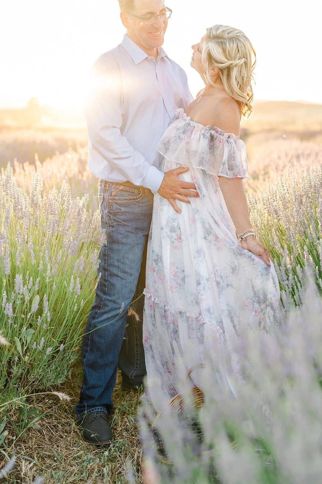 A couple standing closely in a lavender field during sunset, with the man smiling at the woman who is holding her dress, both dressed casually.