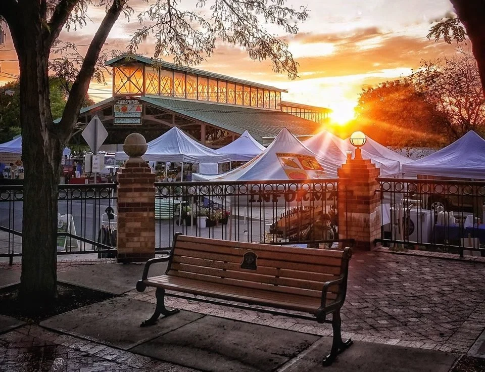 Sunset over outdoor market with white tents, brick fence, and a park bench in foreground.