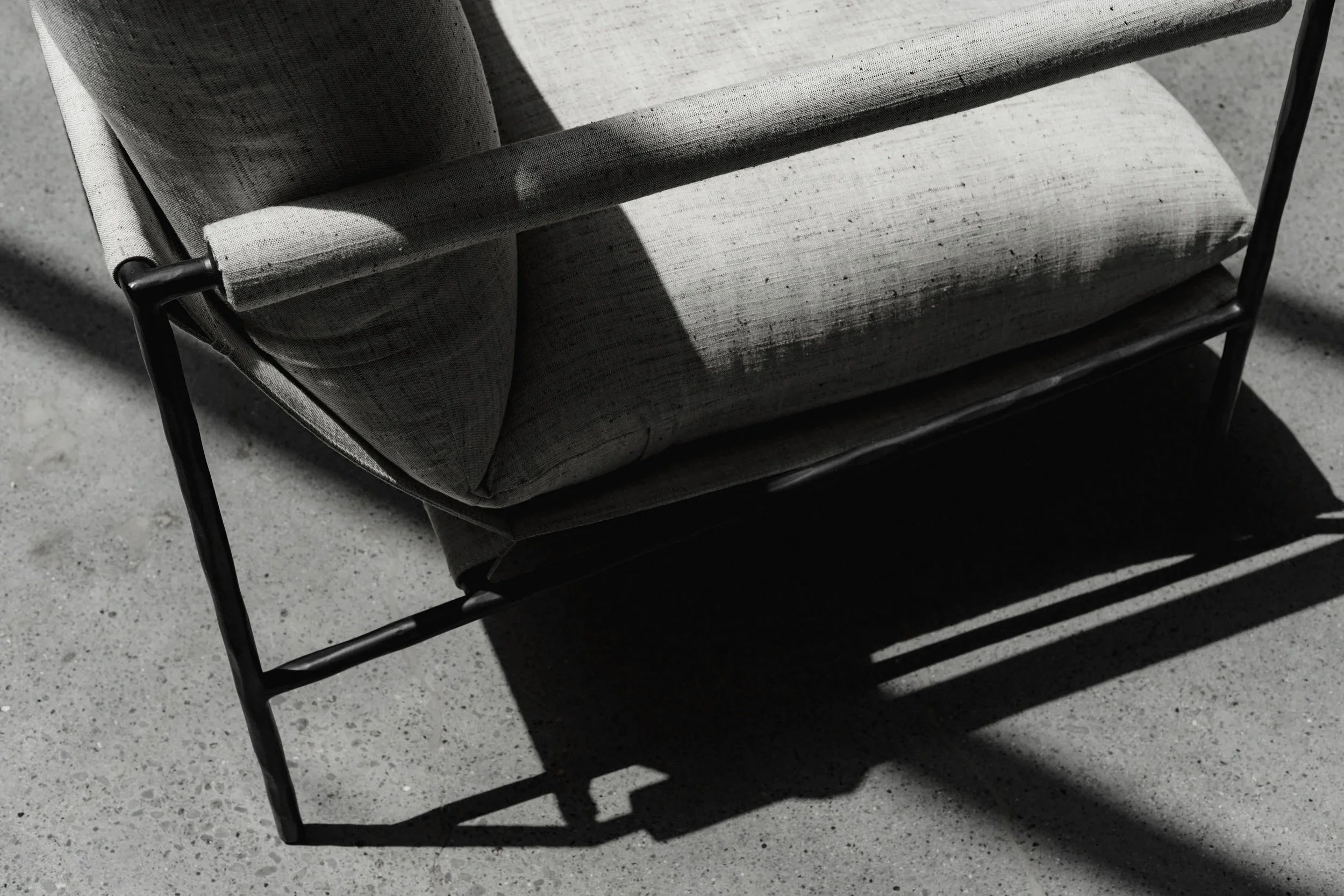 A close-up of a modern outdoor chair with a light grey fabric cushion, metal arms, and shadows cast on a concrete surface.