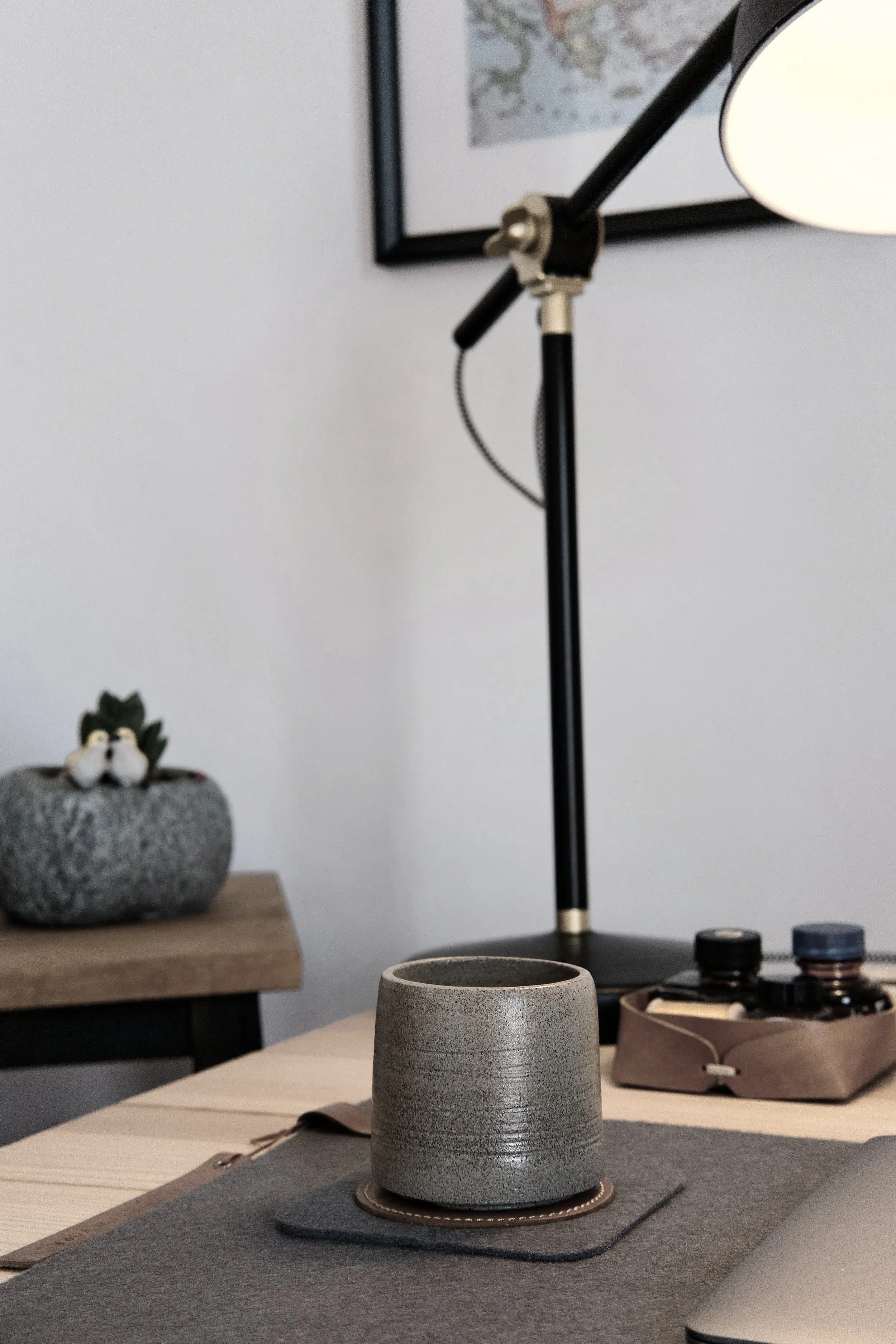 A gray ceramic cup on a wooden table with a black placemat underneath, in a room with decorative objects including a framed picture on the wall, a stone pot, and a tray with bottles, lamps, and other items in the background.