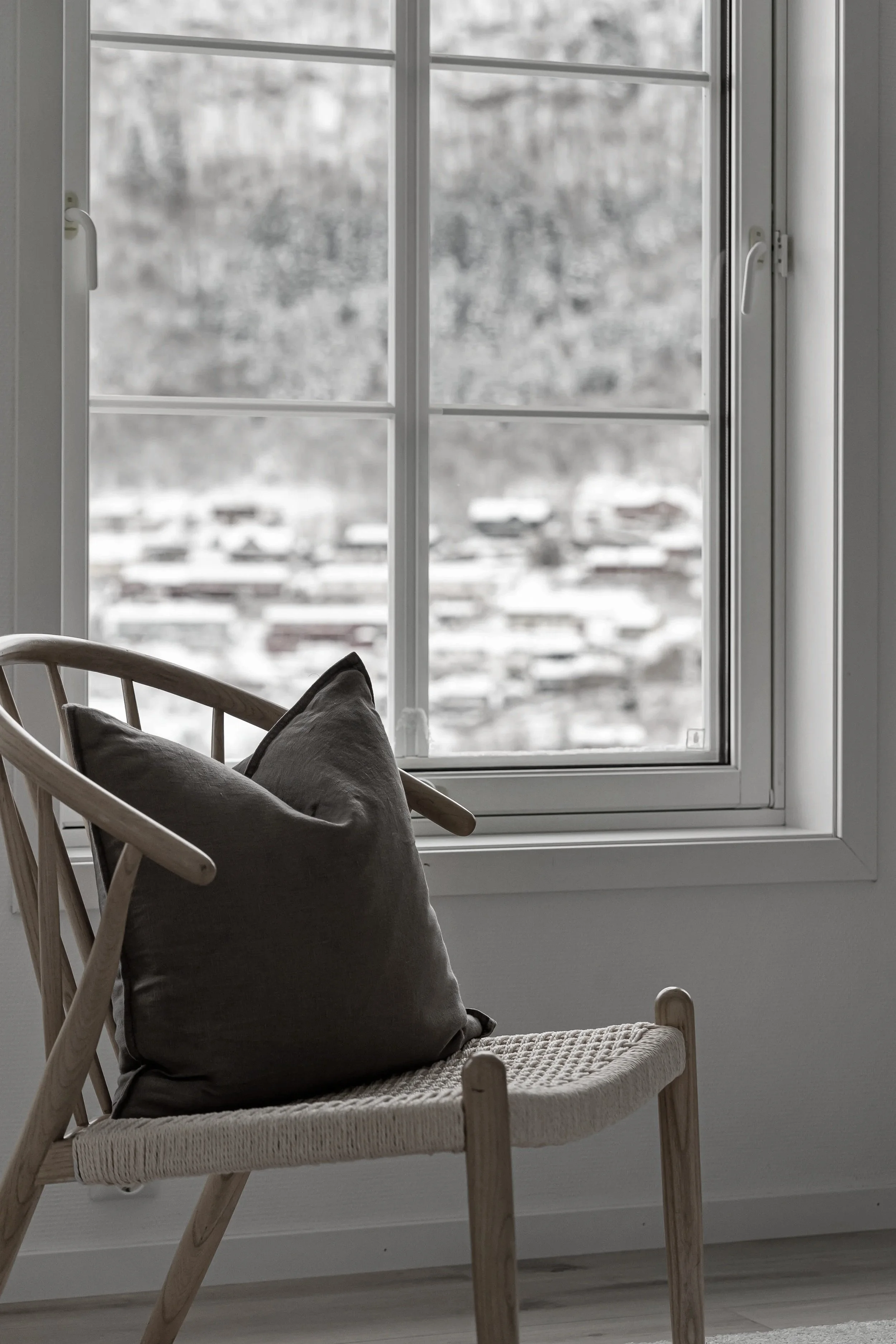 A wooden chair with a cushion and a pillow, positioned in front of a window with white frames showing a snowy landscape outside.