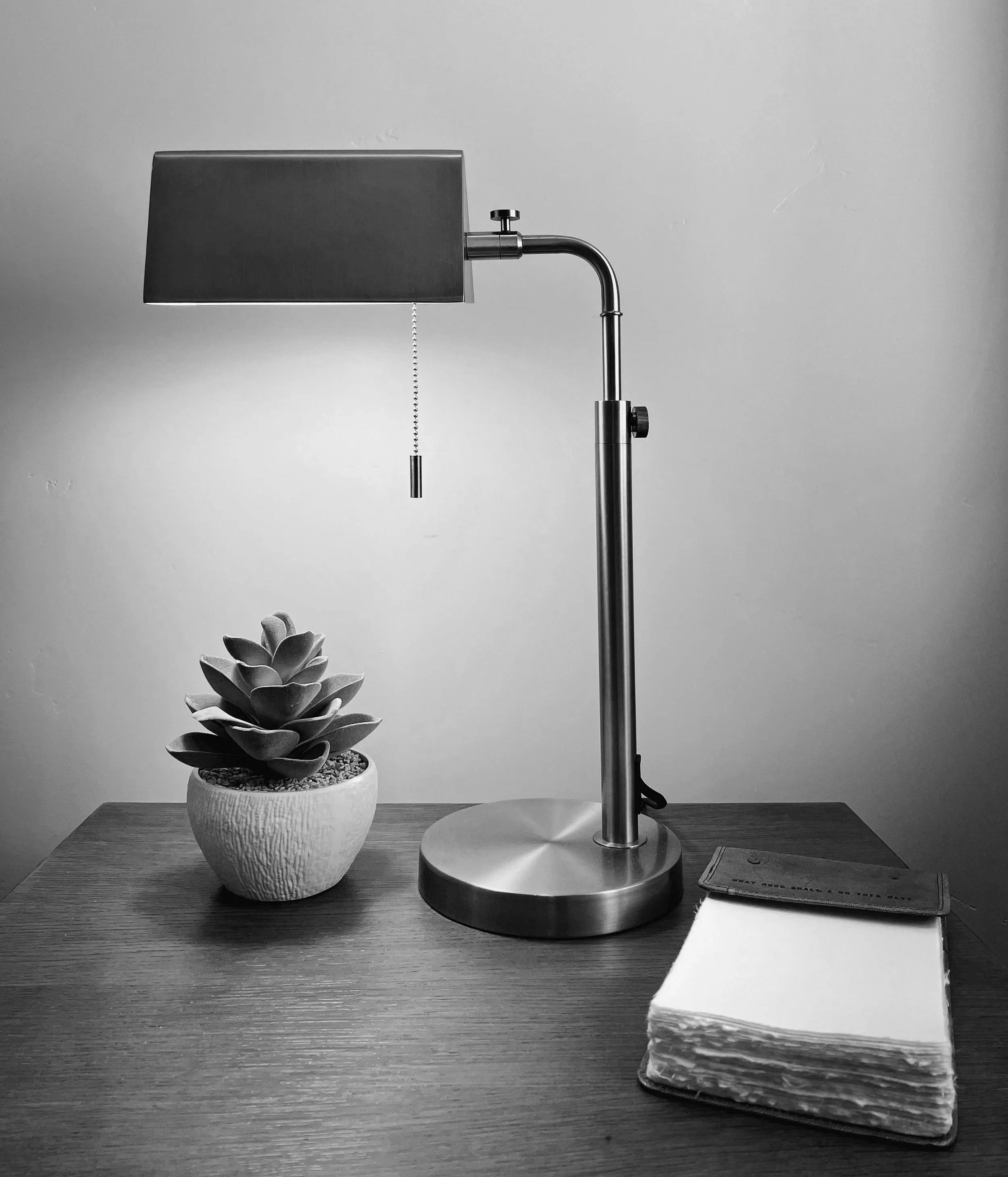 A desk with a potted succulent plant, a modern desk lamp, a closed notebook, and a stack of papers or napkins in black and white.