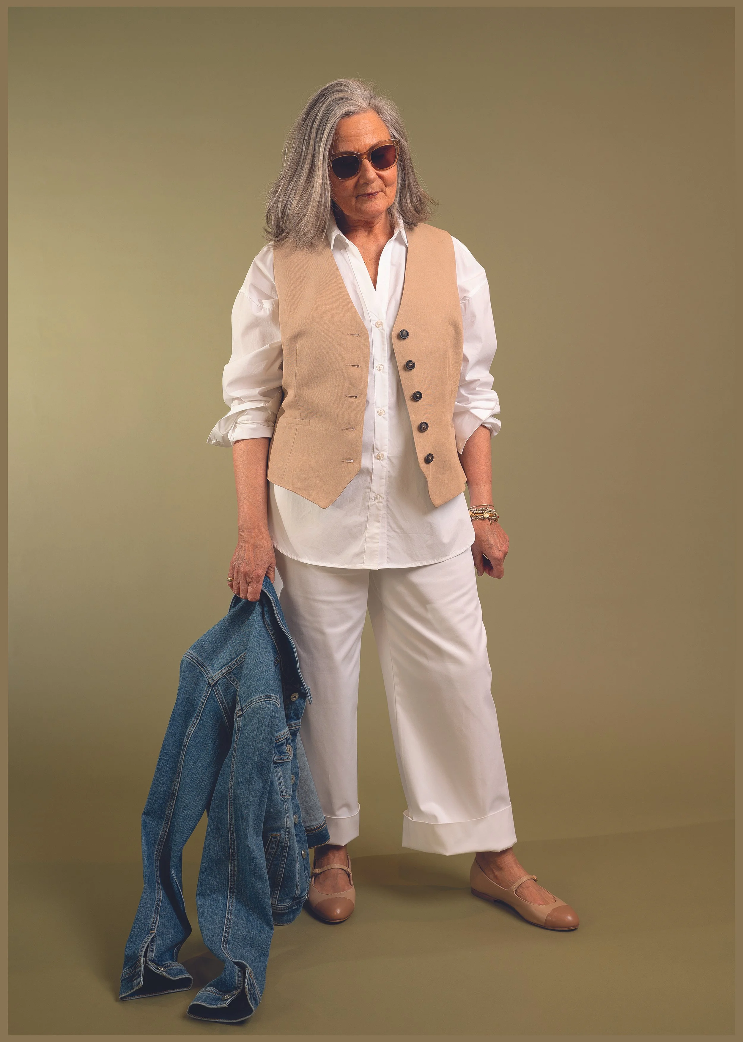 Ältere Frau mit weißem Hemd und beiger Weste, trägt Sonnenbrille, hält eine Jeansjacke in der Hand, Hintergrund in Olivgrün.