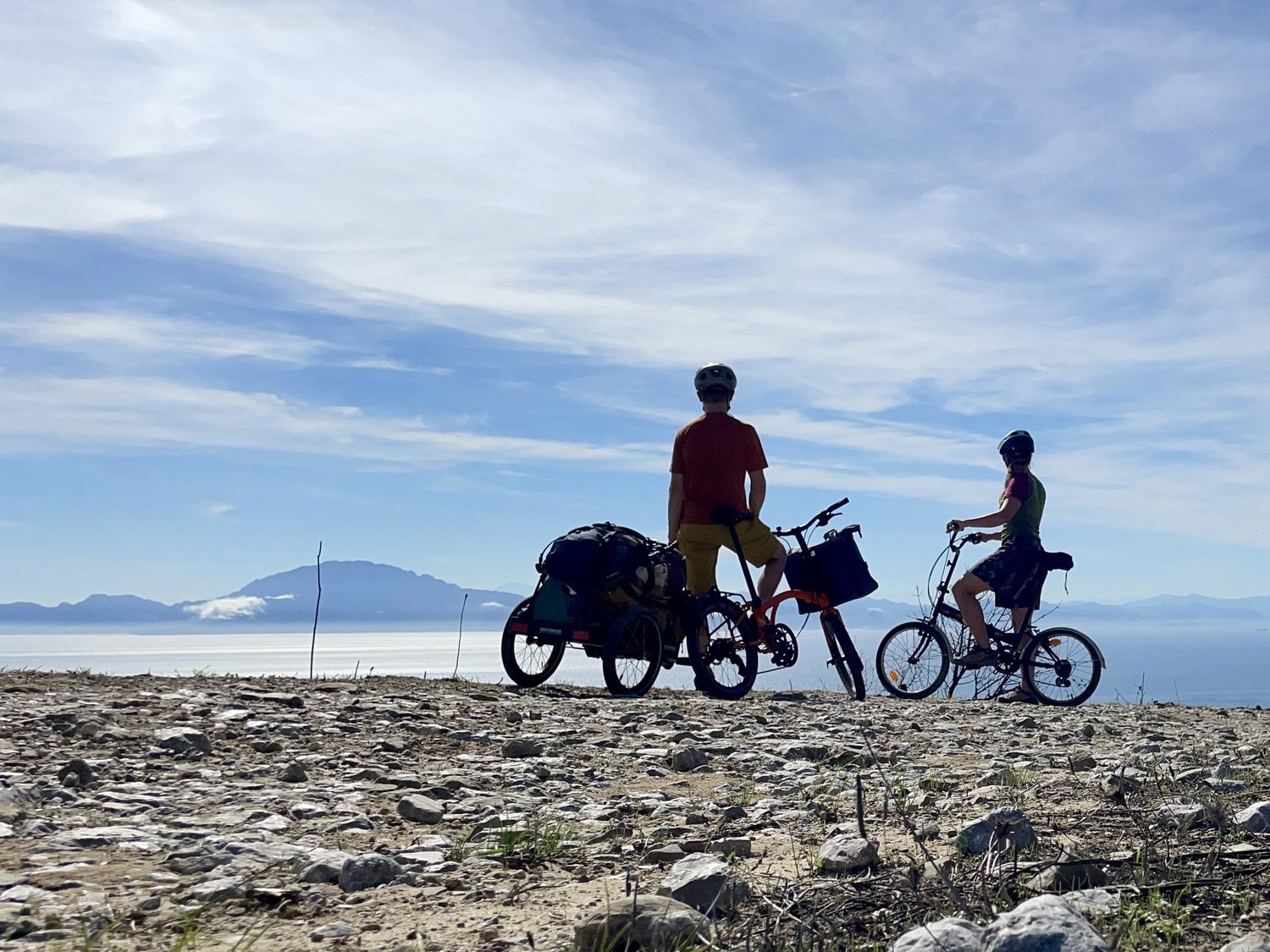 Zwei Radfahrer mit Falträdern und Hinterher Hmini am Meer, im Hintergrund Berge