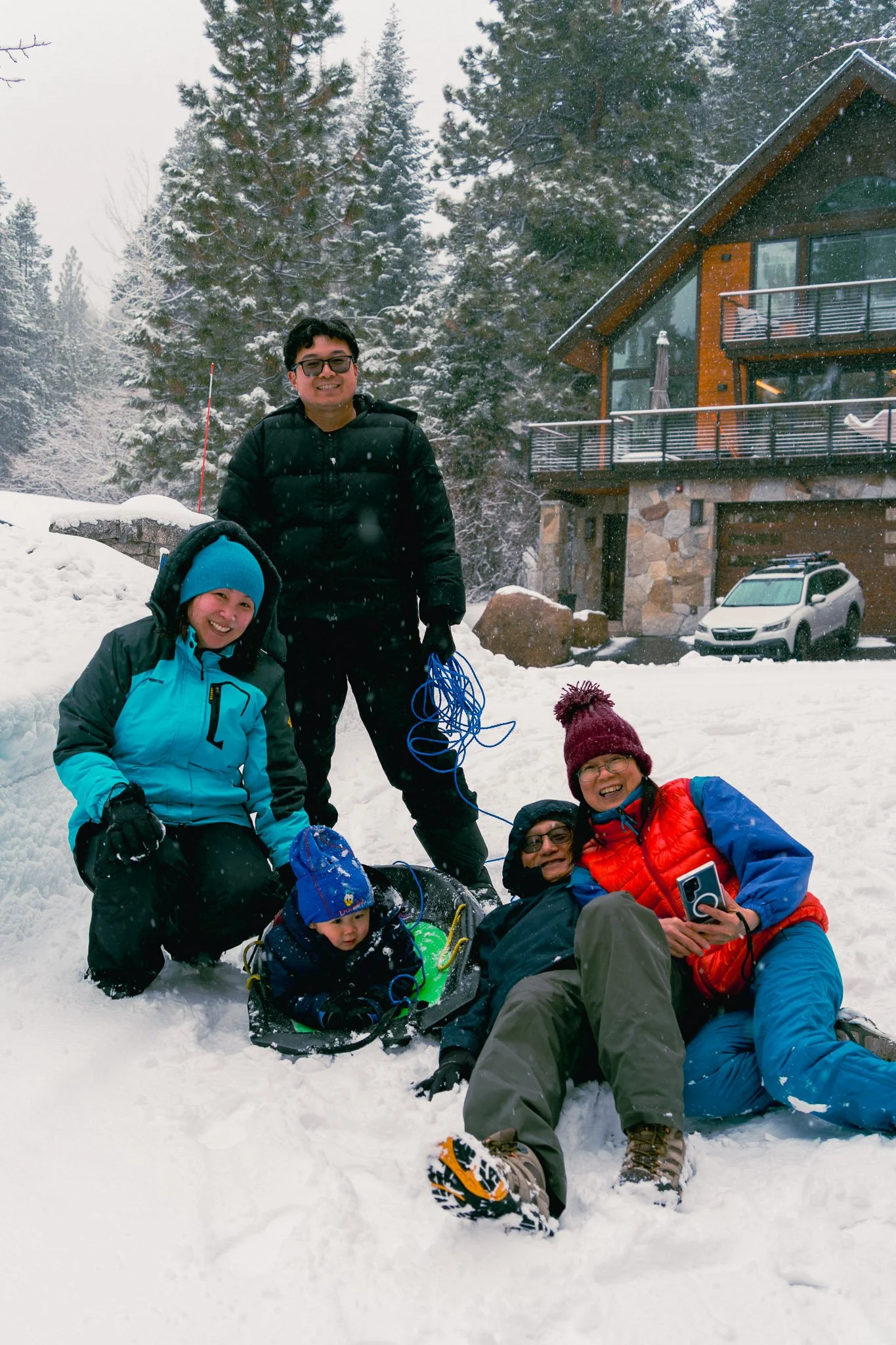 Group of five people in winter clothing enjoying a snowy day outside a house with trees in the background.