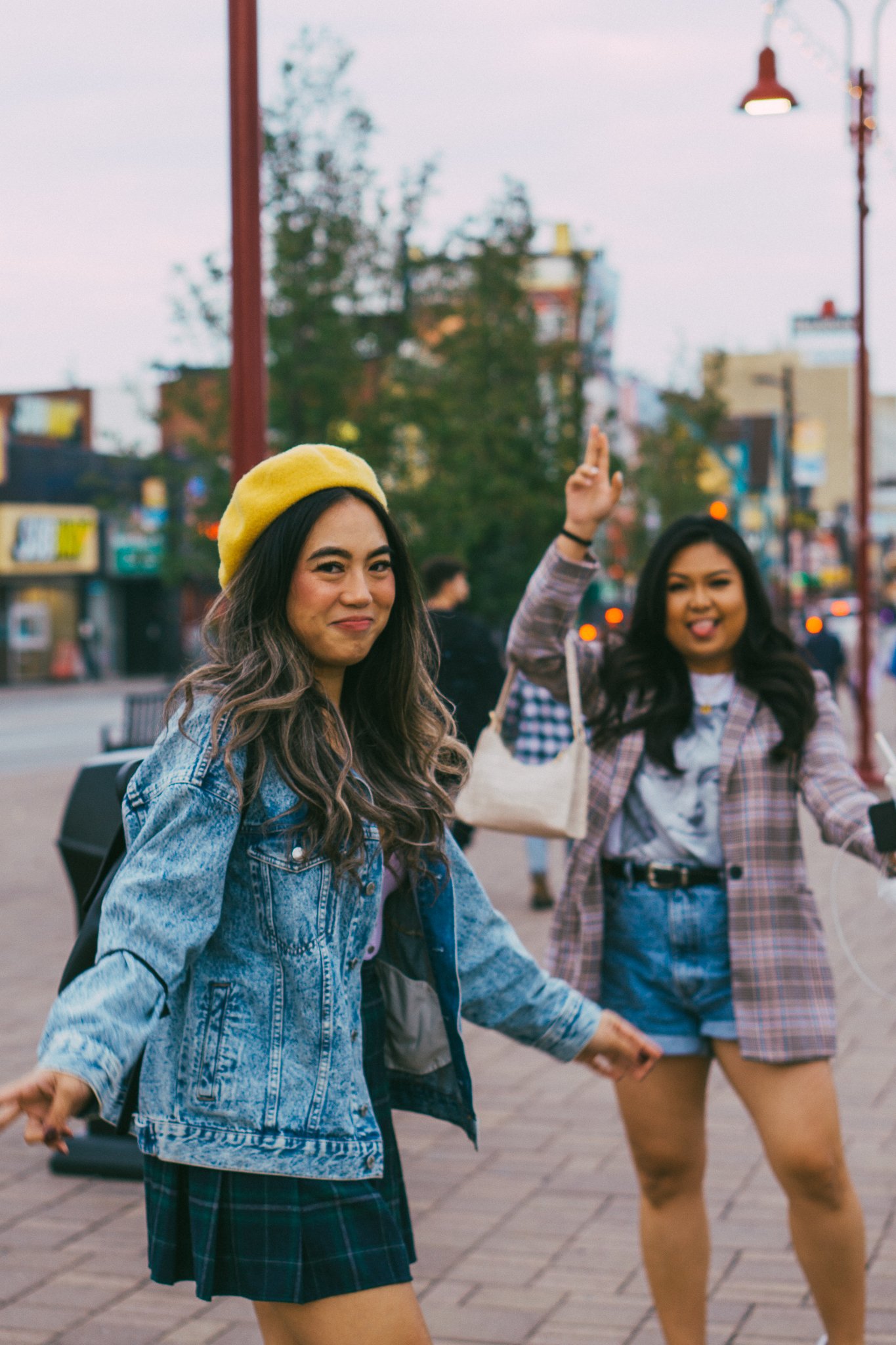 Two young women walking on a city street, one is smiling and wearing a yellow beret and a denim jacket, the other is sticking out her tongue and raising her hand.