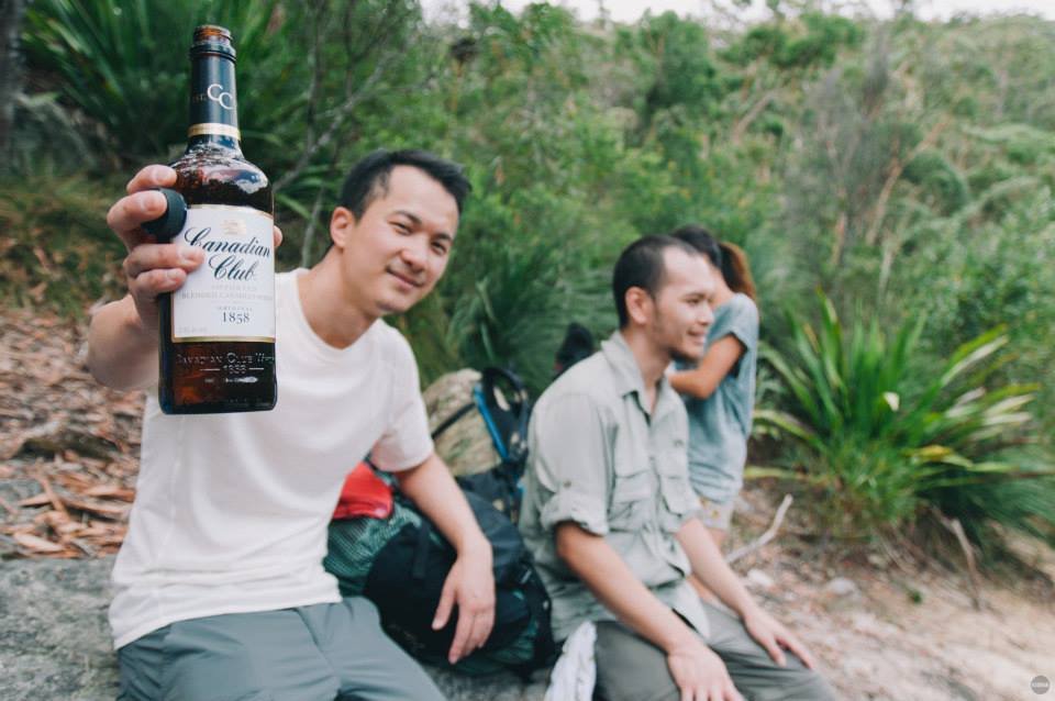 A man holding a bottle of Canadian Club whiskey in a forested outdoor setting, with two other people sitting nearby and smiling.