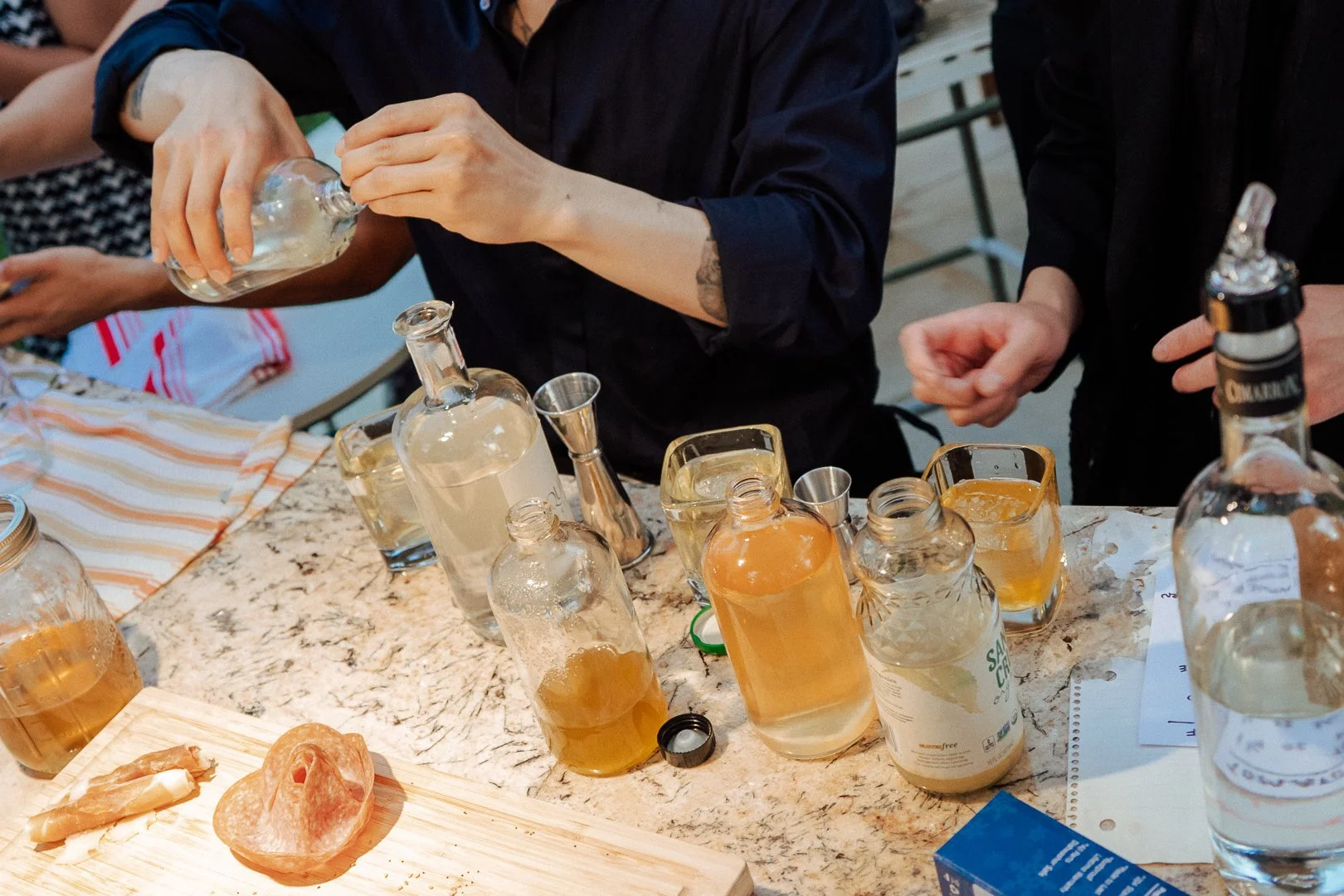 People pouring alcohol into glass bottles on a kitchen countertop with various bottles and jars filled with liquids.