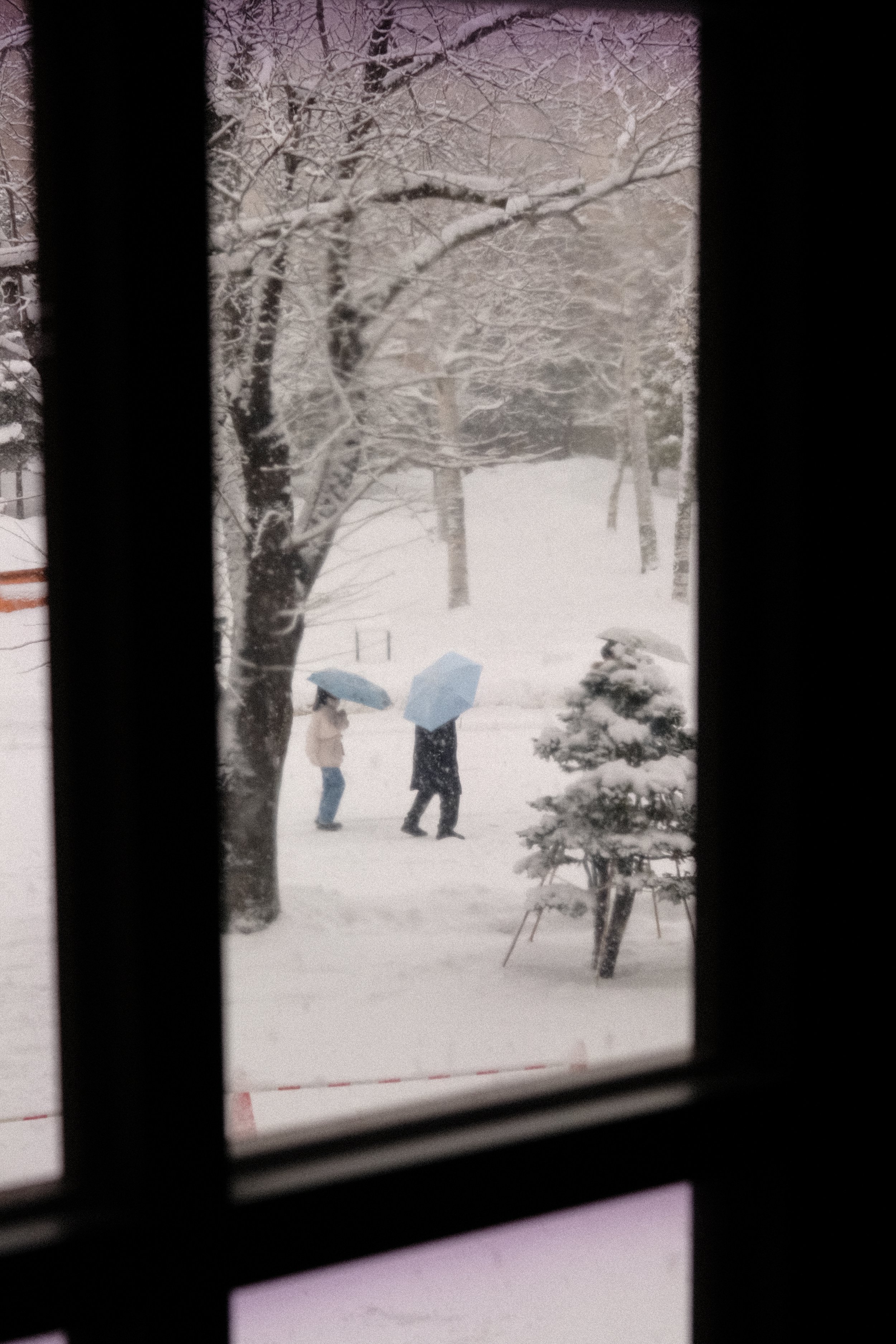 Snow-covered park seen through a window with two people walking with umbrellas.