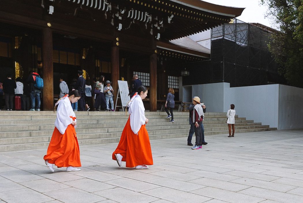 Two women dressed in traditional Japanese shrine maidens' attire walking in front of a temple entrance with many visitors and a few children.
