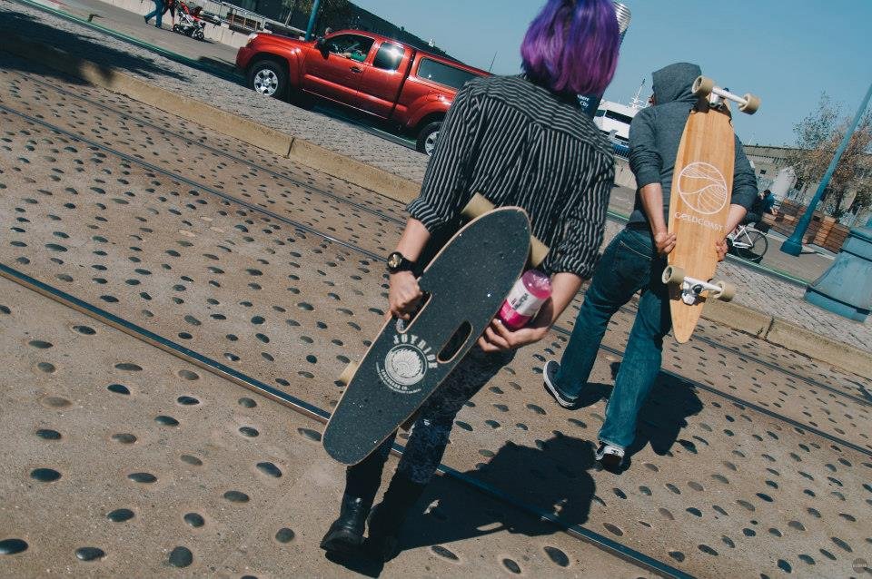 Two skateboarders, one with purple hair and the other with a gray hoodie, walking on a sidewalk near train tracks, holding skateboards, with a red pickup truck and people in the background.