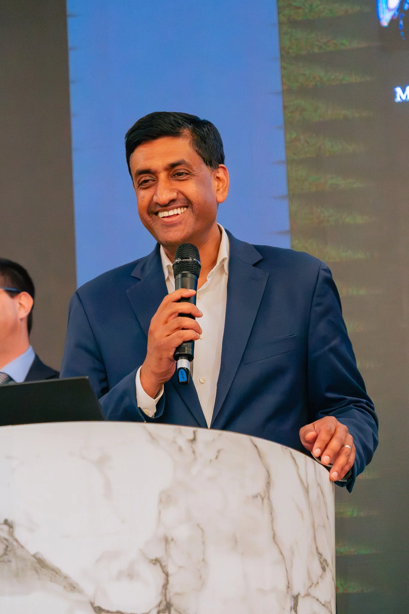 A man in a navy blue suit and white shirt speaking into a microphone at a podium during a conference or event.
