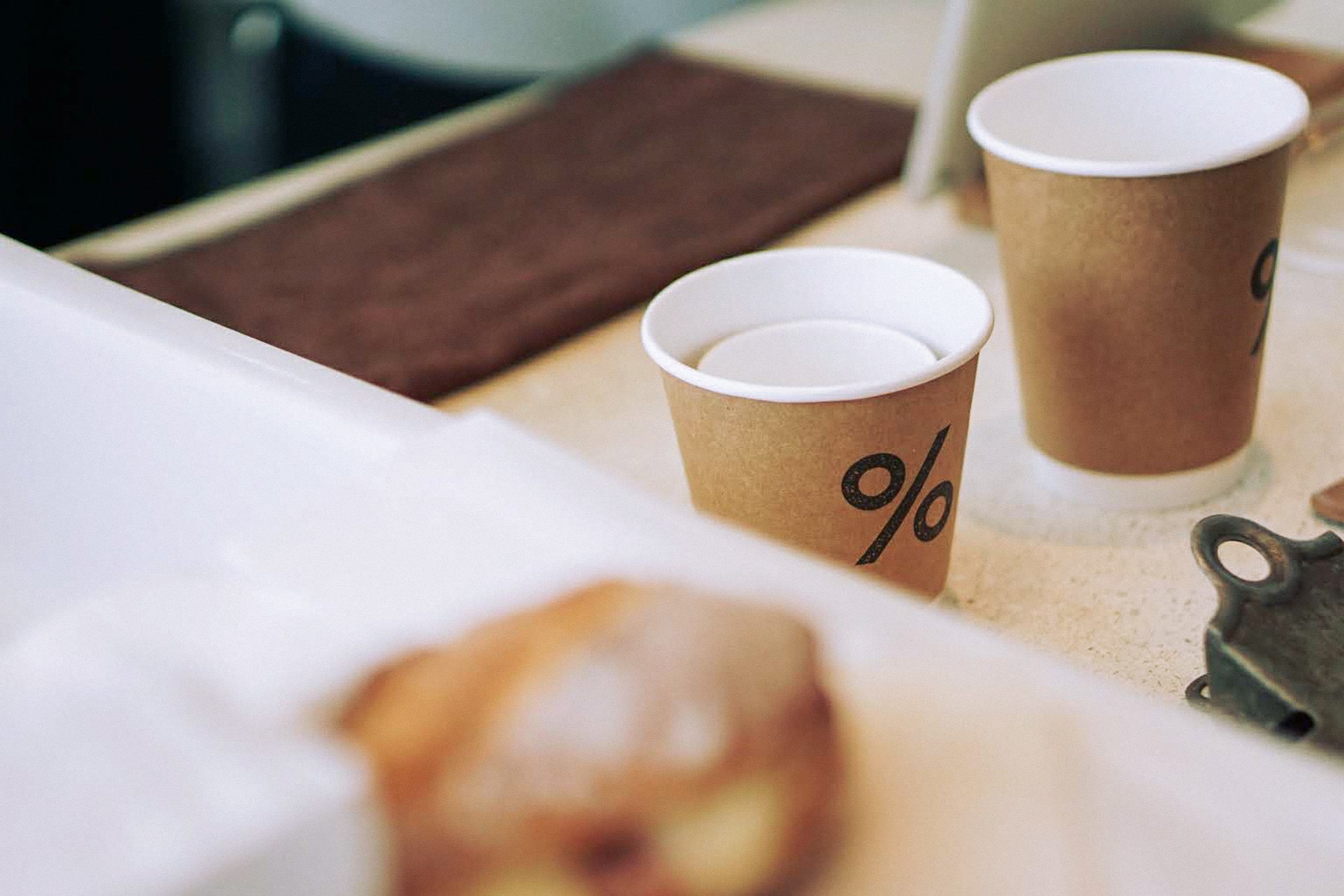 Two disposable coffee cups with a percent symbol printed on them, placed on a light-colored surface, with one cup in the foreground and another in the background.