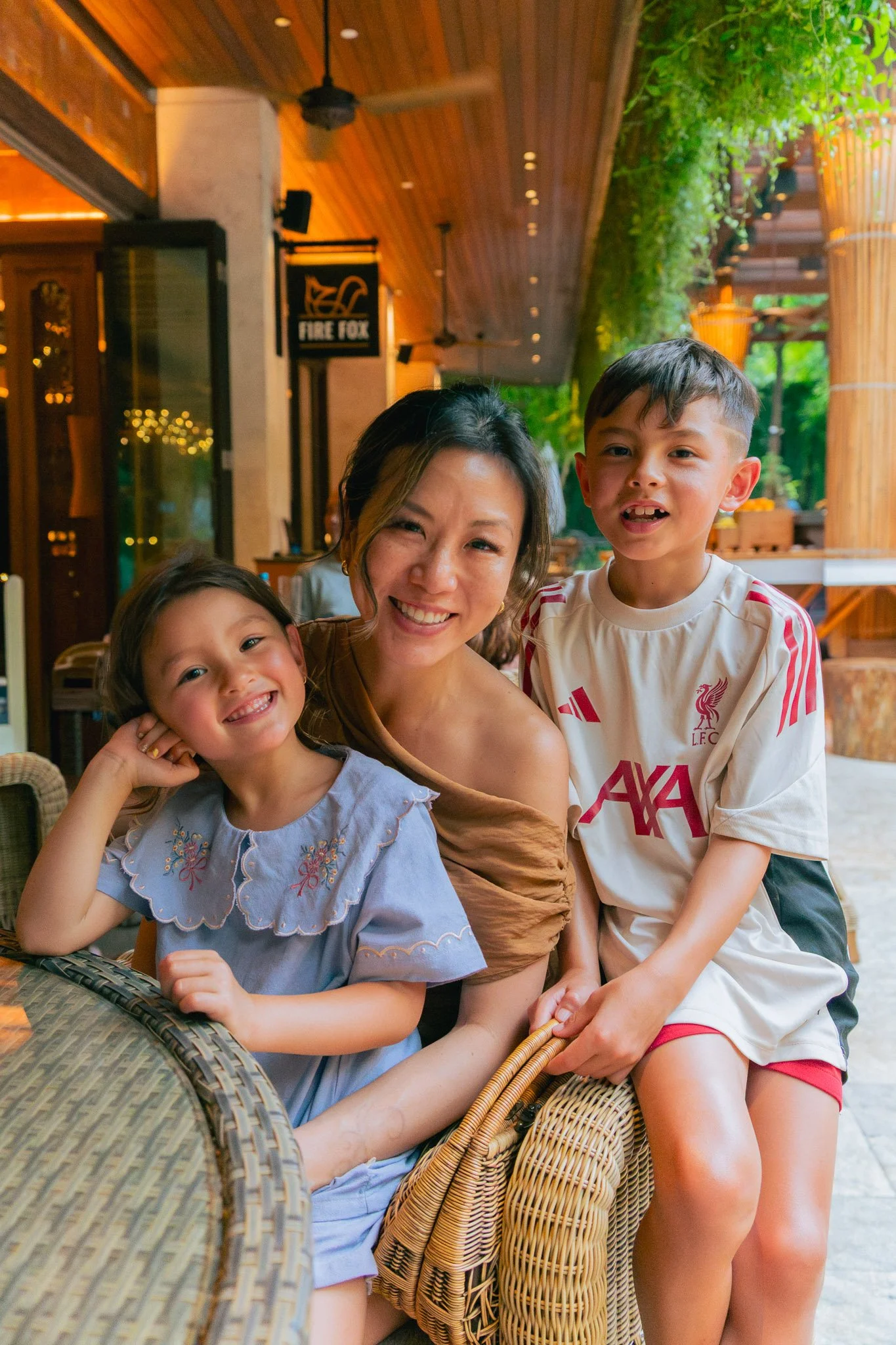 A woman and two children smiling at a restaurant with wooden interior and green plants.