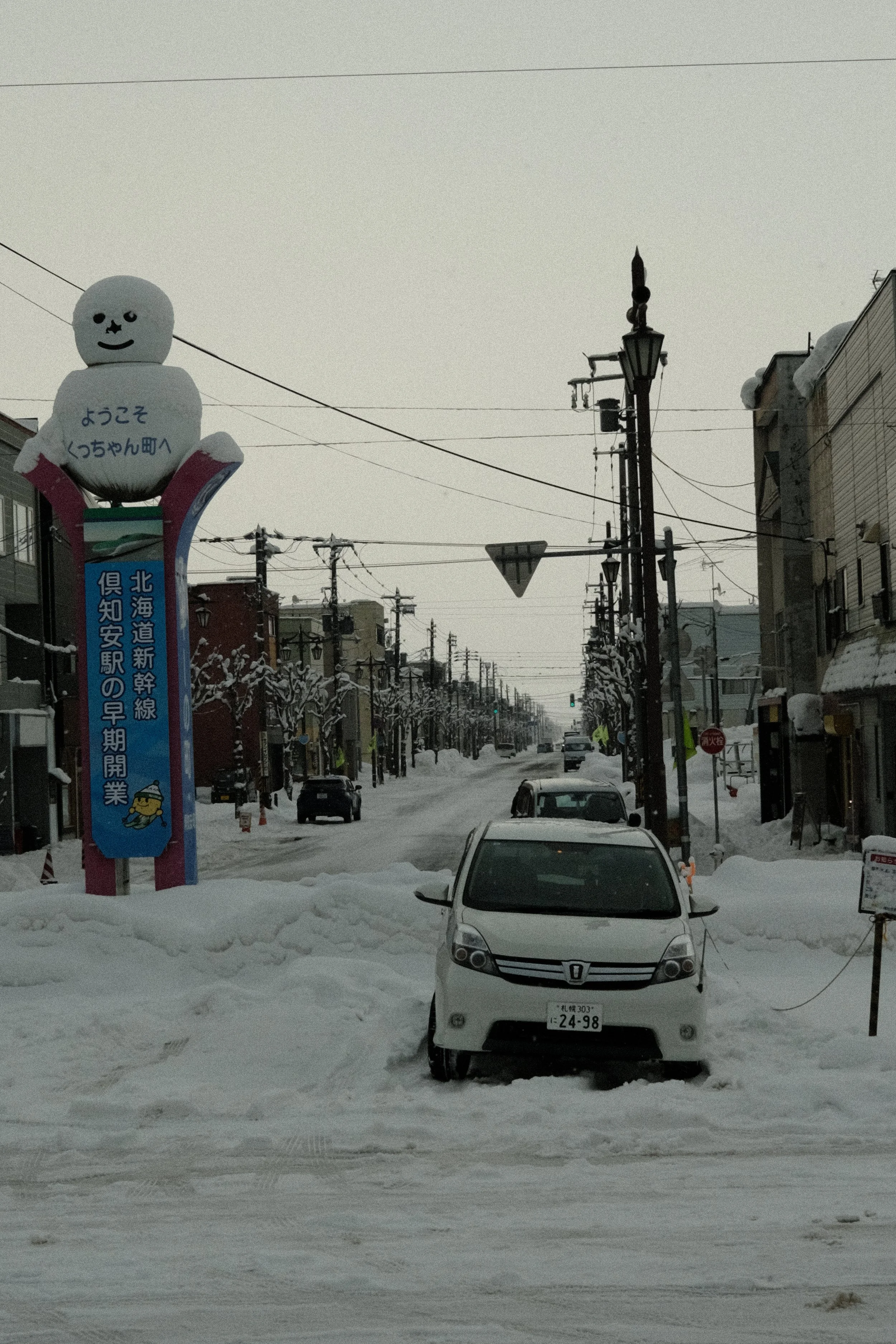 Snow-covered street in a small town with parked cars and buildings on either side. A large cartoon snowman sculpture with a smiling face and Japanese writing stands near a colorful sign on the left. Power lines run overhead.