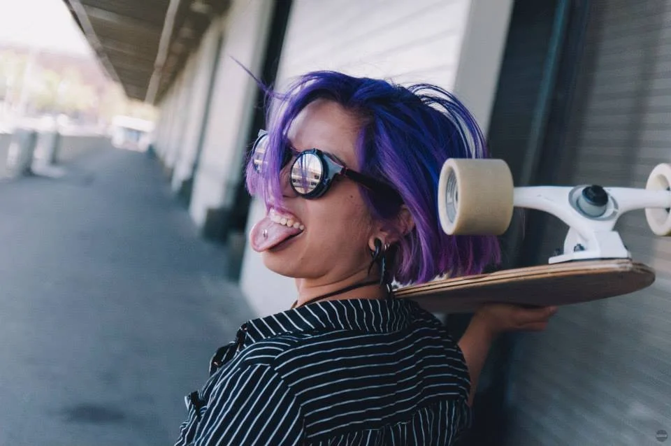 Young woman with purple hair and sunglasses sticking out her tongue, holding a skateboard over her shoulder, standing outdoors in front of a building.