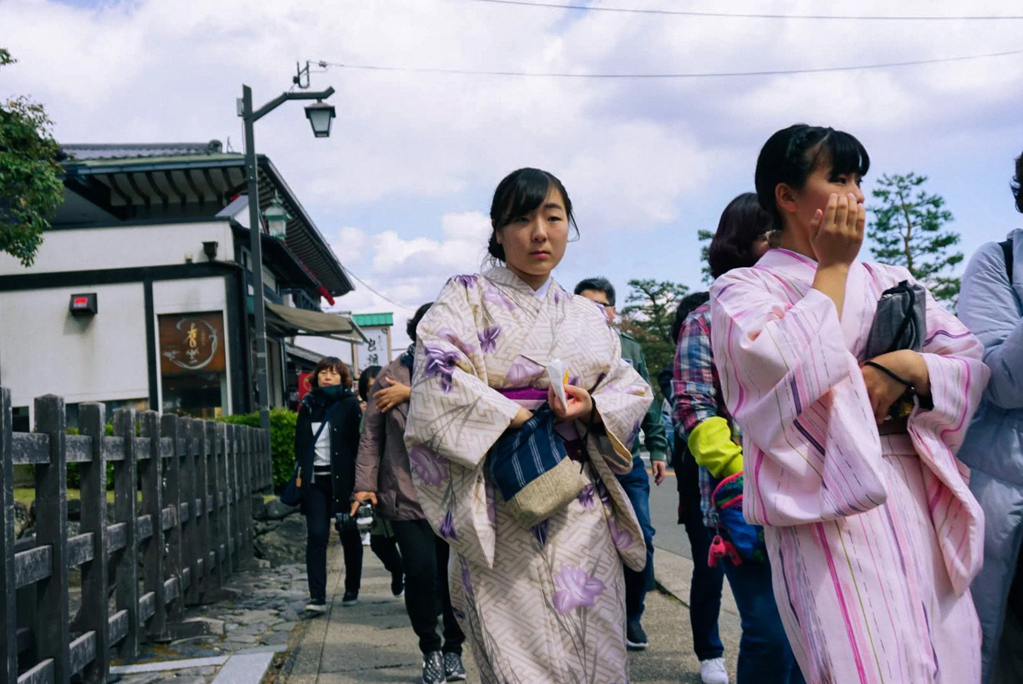 Group of people, including women in kimonos, walking outdoors on a street with buildings and trees in the background.