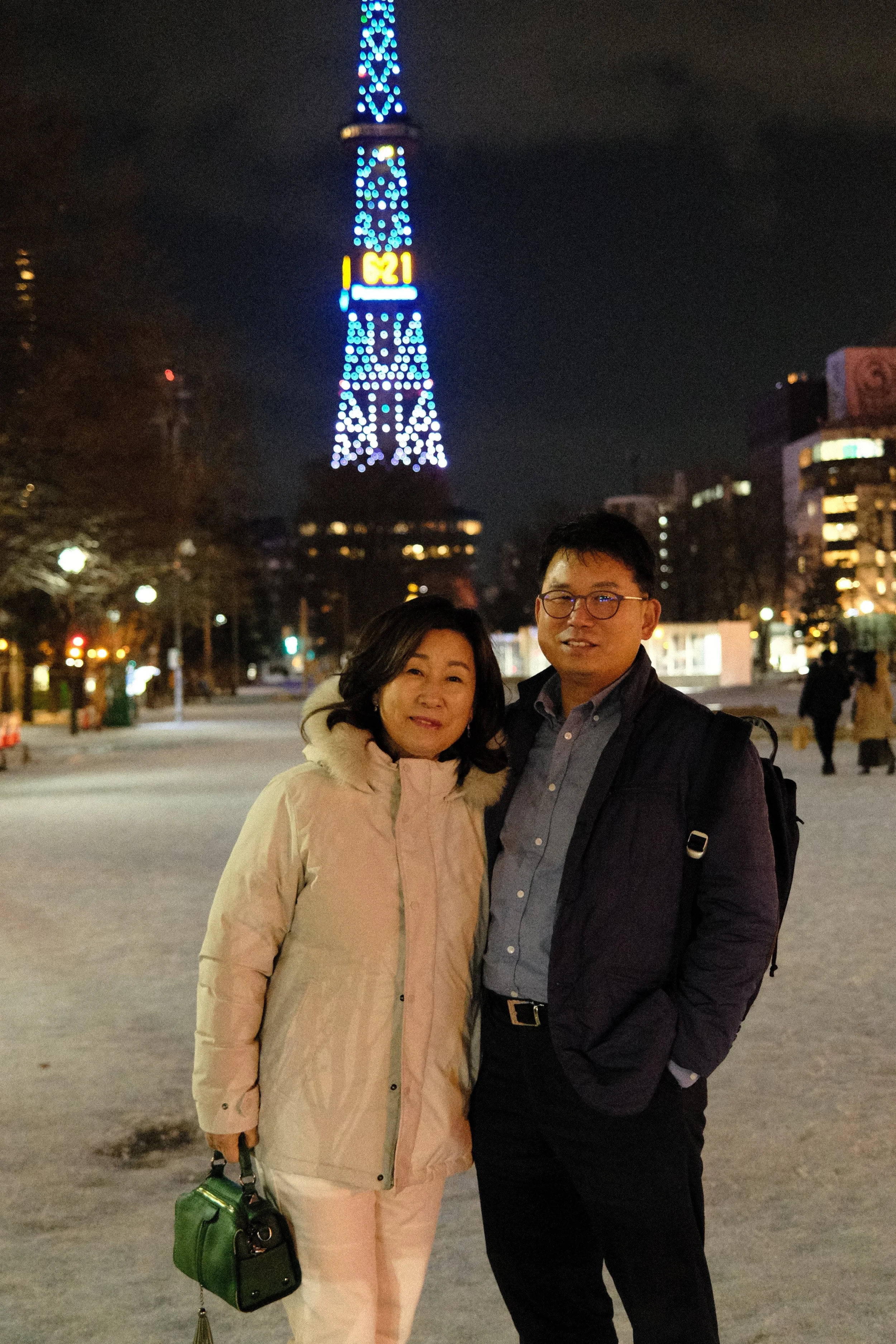 A woman and a man standing together outdoors at night in a snowy area, with a tall illuminated tower with lights and digital displays in the background.