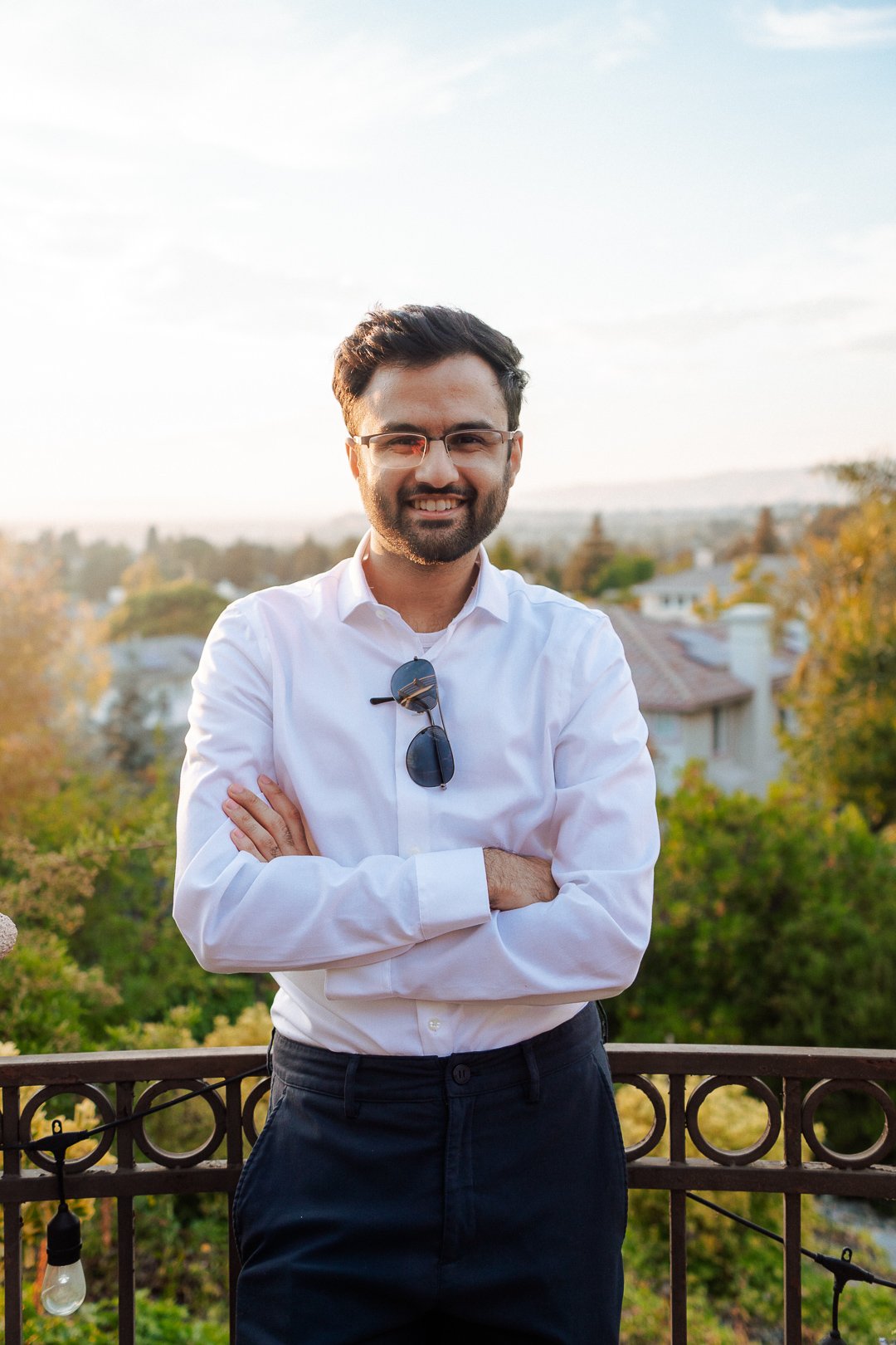 A man with glasses and a beard wearing a white shirt and black pants standing outdoors on a balcony with arms crossed, smiling at the camera, with a backdrop of trees and houses under a clear sky.