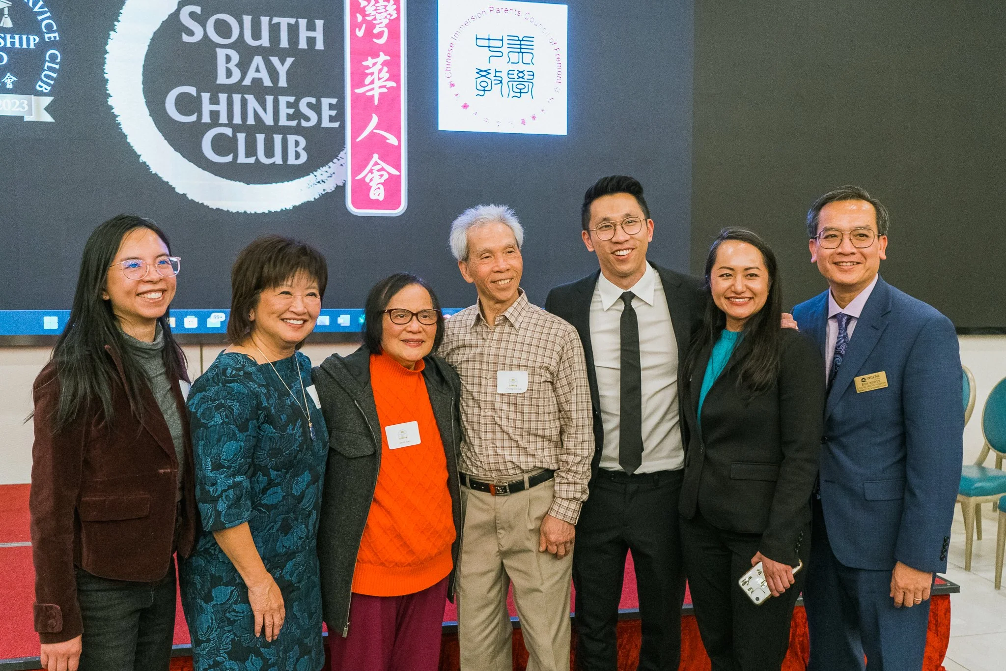 Group of seven people standing together smiling at a social event, with a sign that reads "South Bay Chinese Club" in the background.