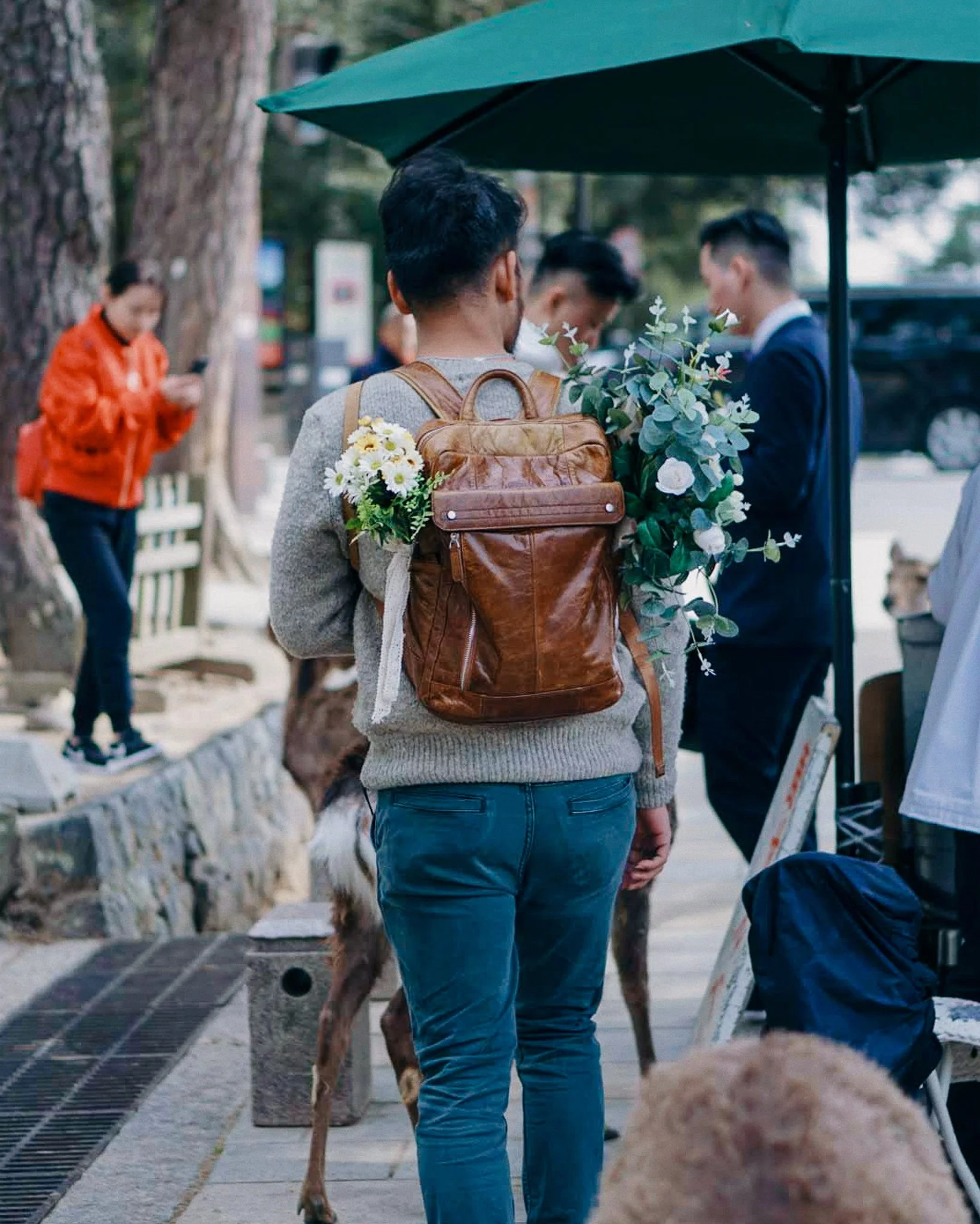Person with a brown backpack decorated with flowers, standing outdoors near a dog, with a group of people and a dog on a leash nearby.