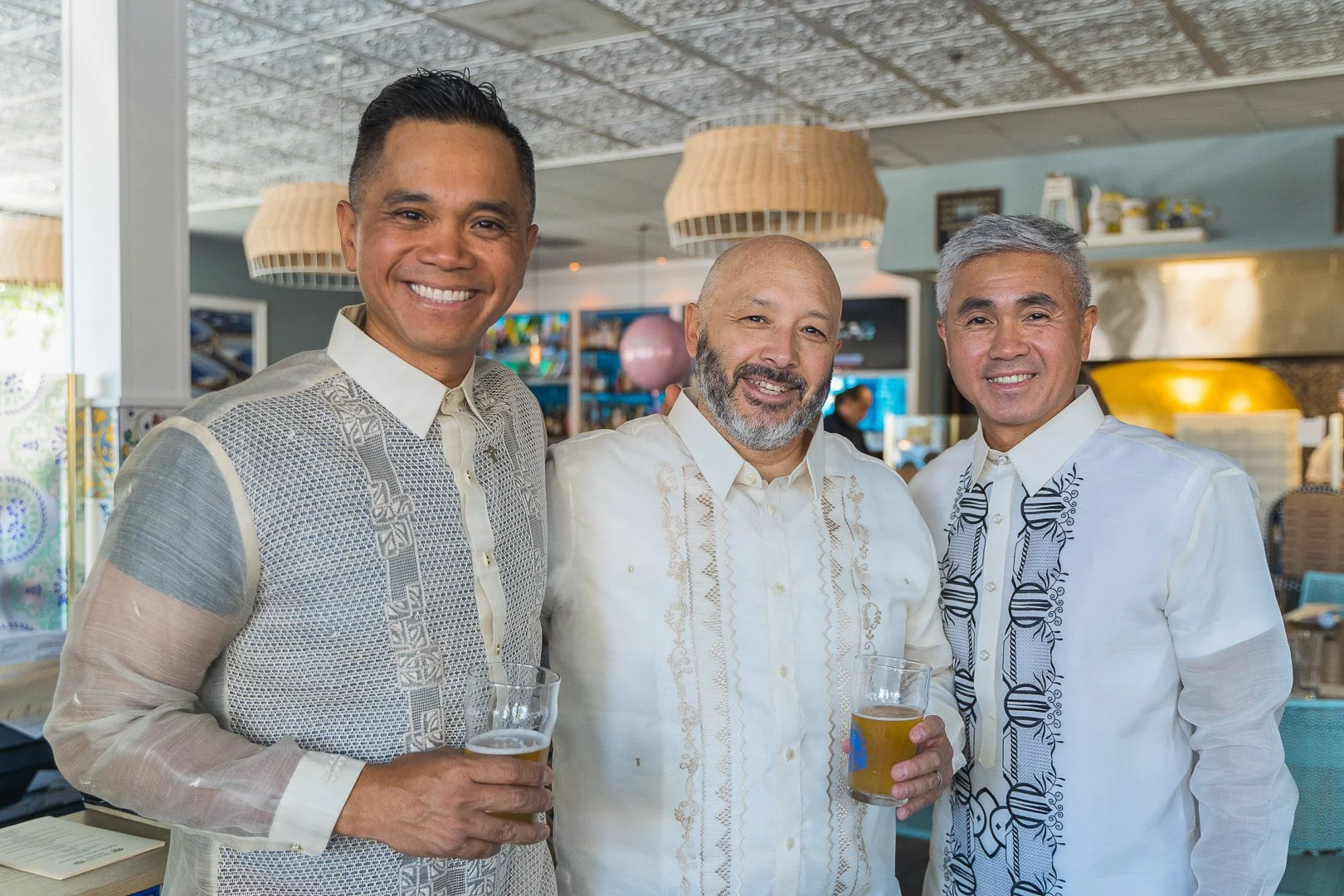 Three men in white shirts smiling and holding drinks in a tropical-themed restaurant or bar.