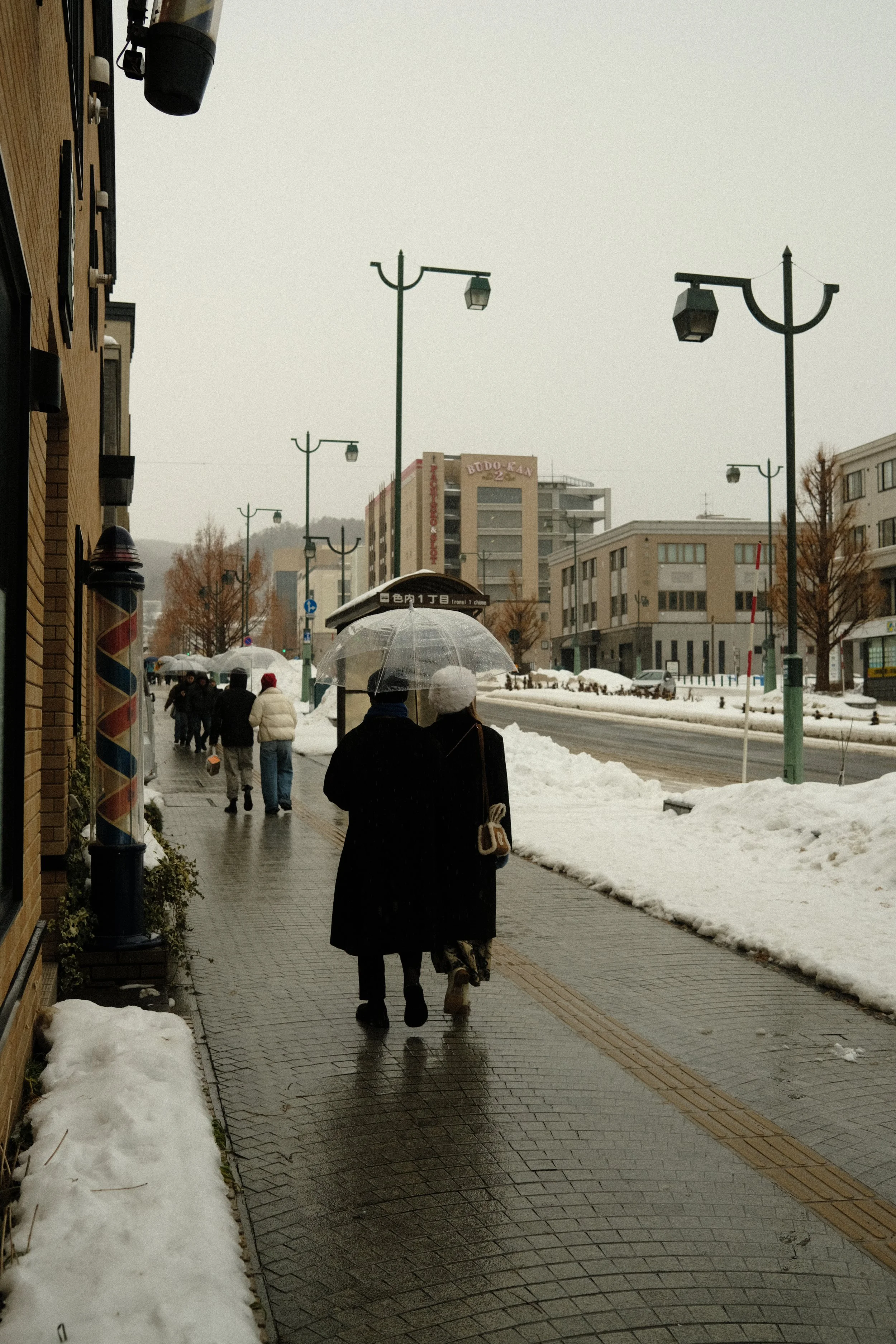 People walking on a city sidewalk with snow, holding umbrellas, and dressed in winter clothing.