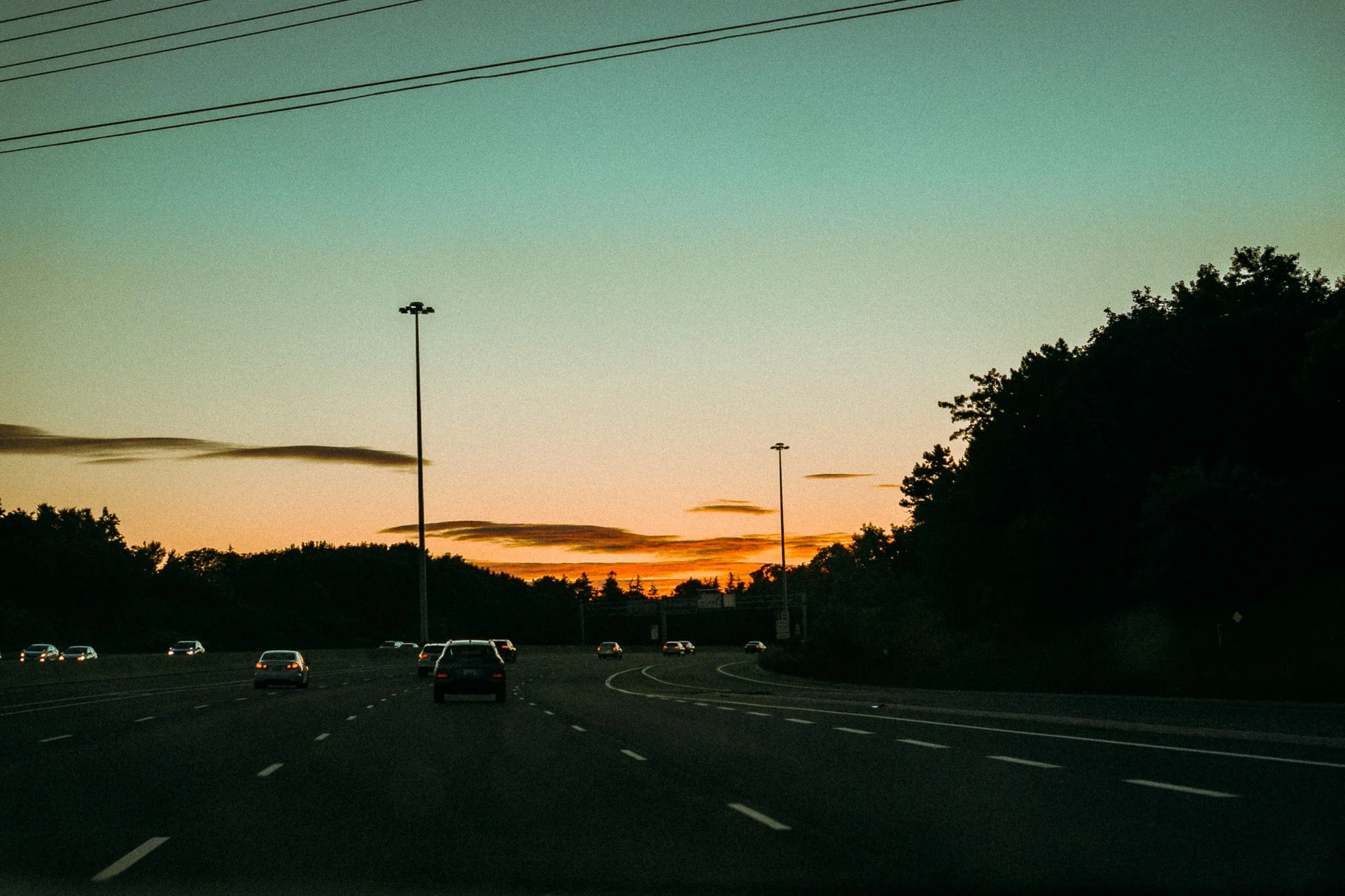 A highway at sunset with cars driving in multiple lanes, tall streetlights, and trees on the right side, with a colorful sky and clouds in the background.