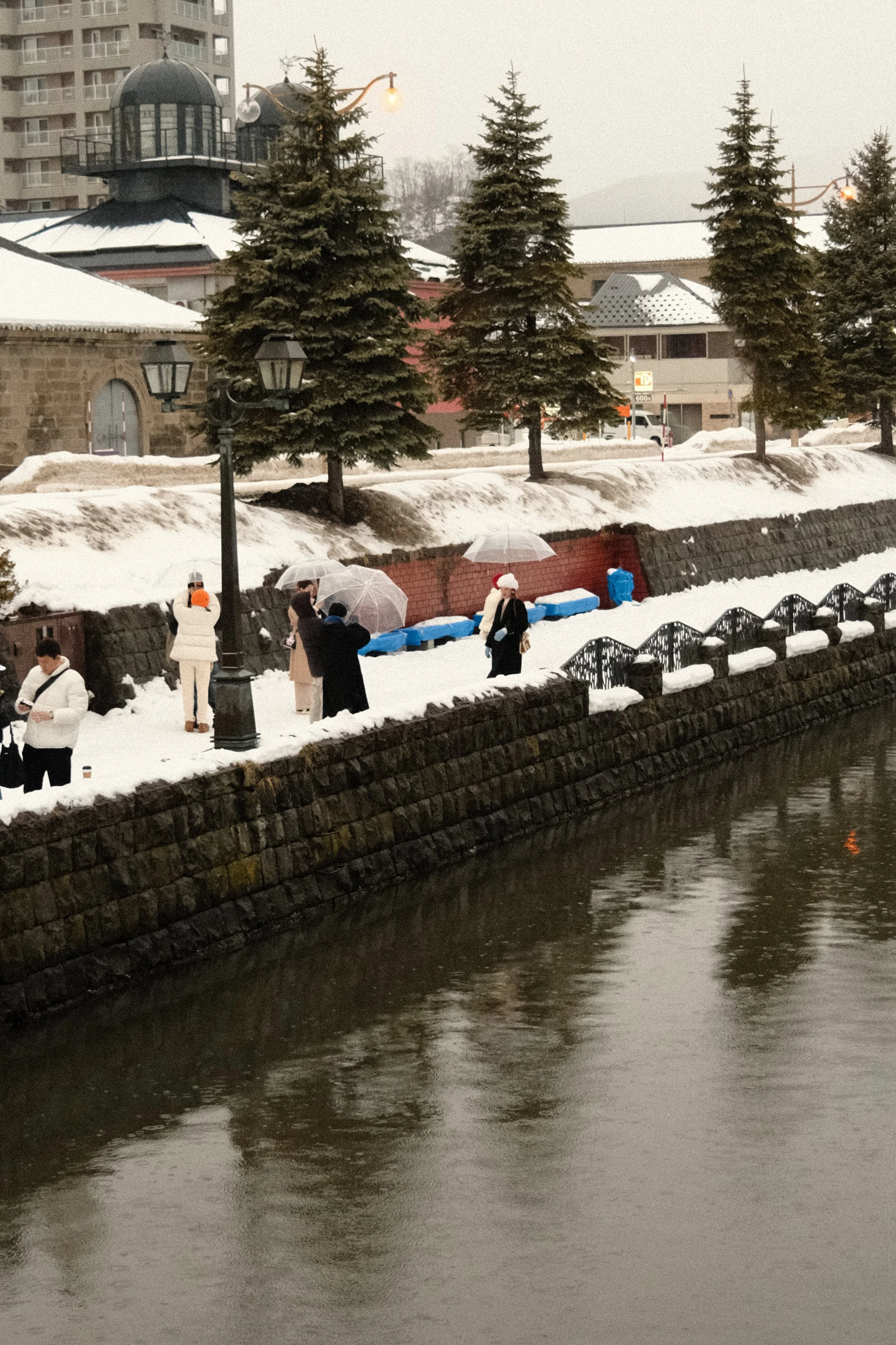 People walking along a snowy riverbank with umbrellas, some wearing warm clothing, near tall evergreen trees and old-fashioned street lamps, with buildings and a snow-covered landscape in the background.