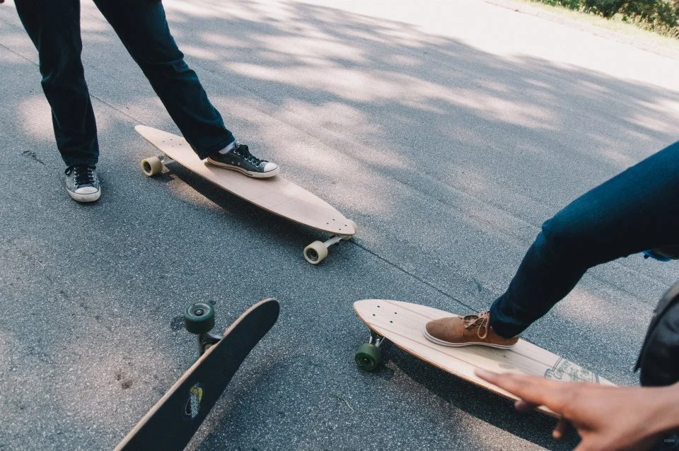 Two people standing on skateboards on a paved road, one wearing blue jeans and black sneakers, the other wearing brown shoes.