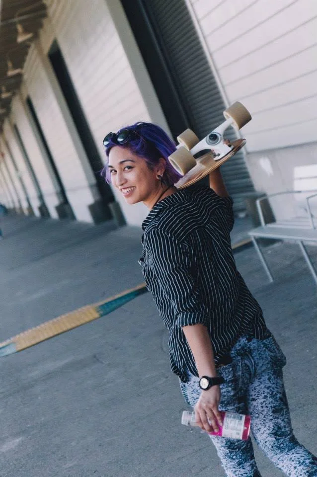 A woman with purple and black hair, wearing sunglasses on her head, a black and white striped shirt, and patterned pants, is holding a skateboard over her shoulder and a drink in her hand, smiling and standing outdoors near a building.
