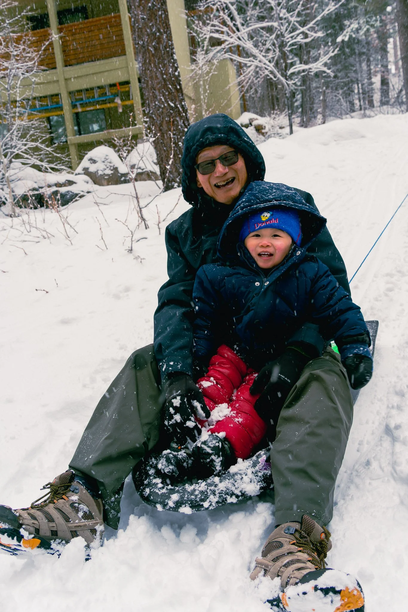 A man and child sledding in a snowy outdoor setting with snow-covered trees and a building in the background.