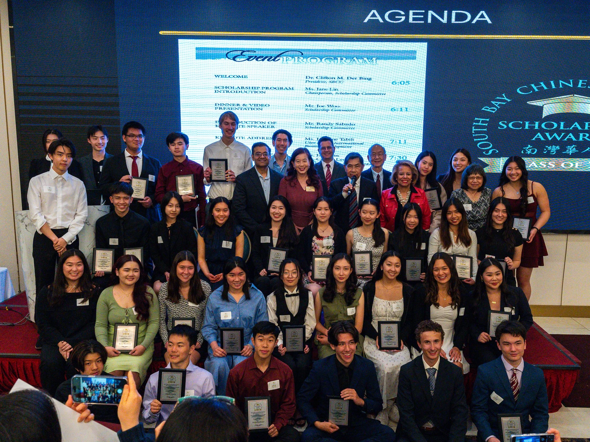Group photo of young scholarship awardees and organizers on stage at South Bay Chinese Scholarship Awards event, holding plaques, with large digital screen displaying event details behind them.