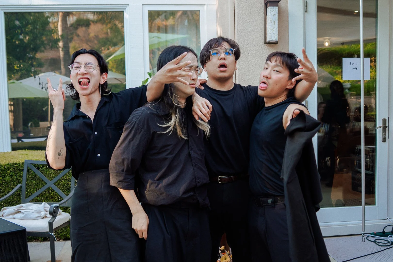 Four friends making funny faces and posing together outside a house, with a glass door and patio furniture in the background.