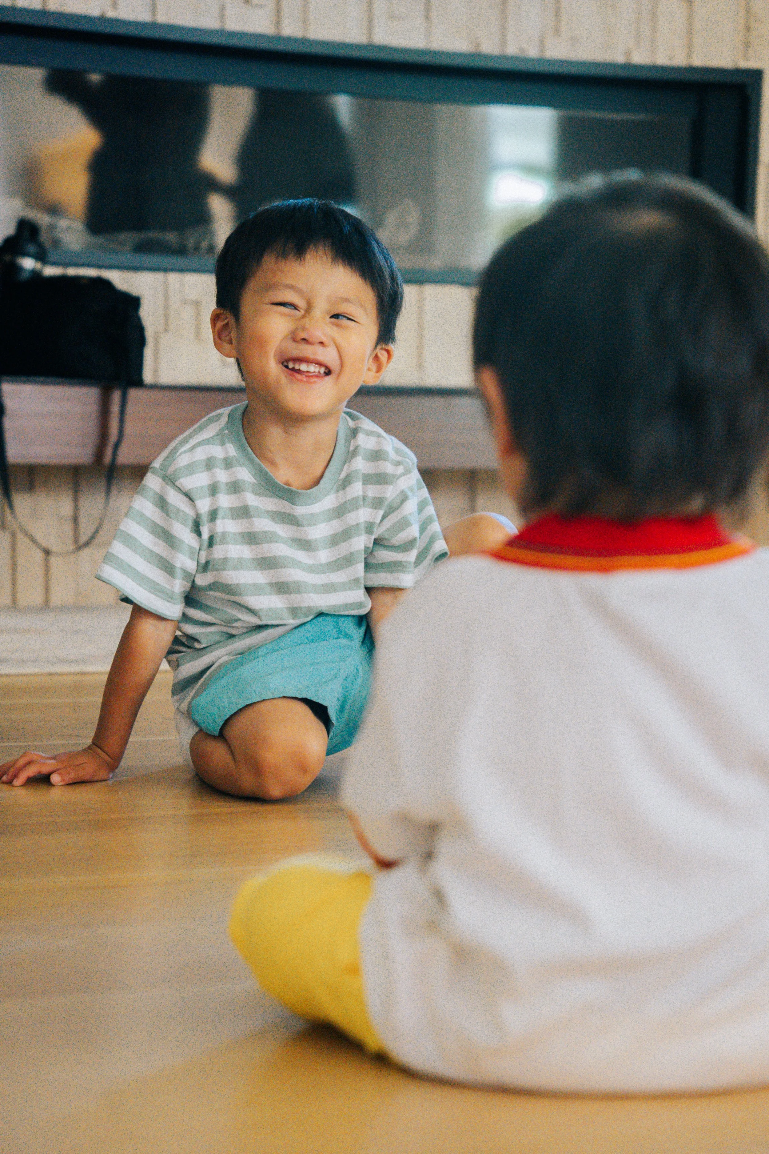 Two children playing on the floor indoors, one smiling and laughing while kneeling and the other sitting with their back to the camera.